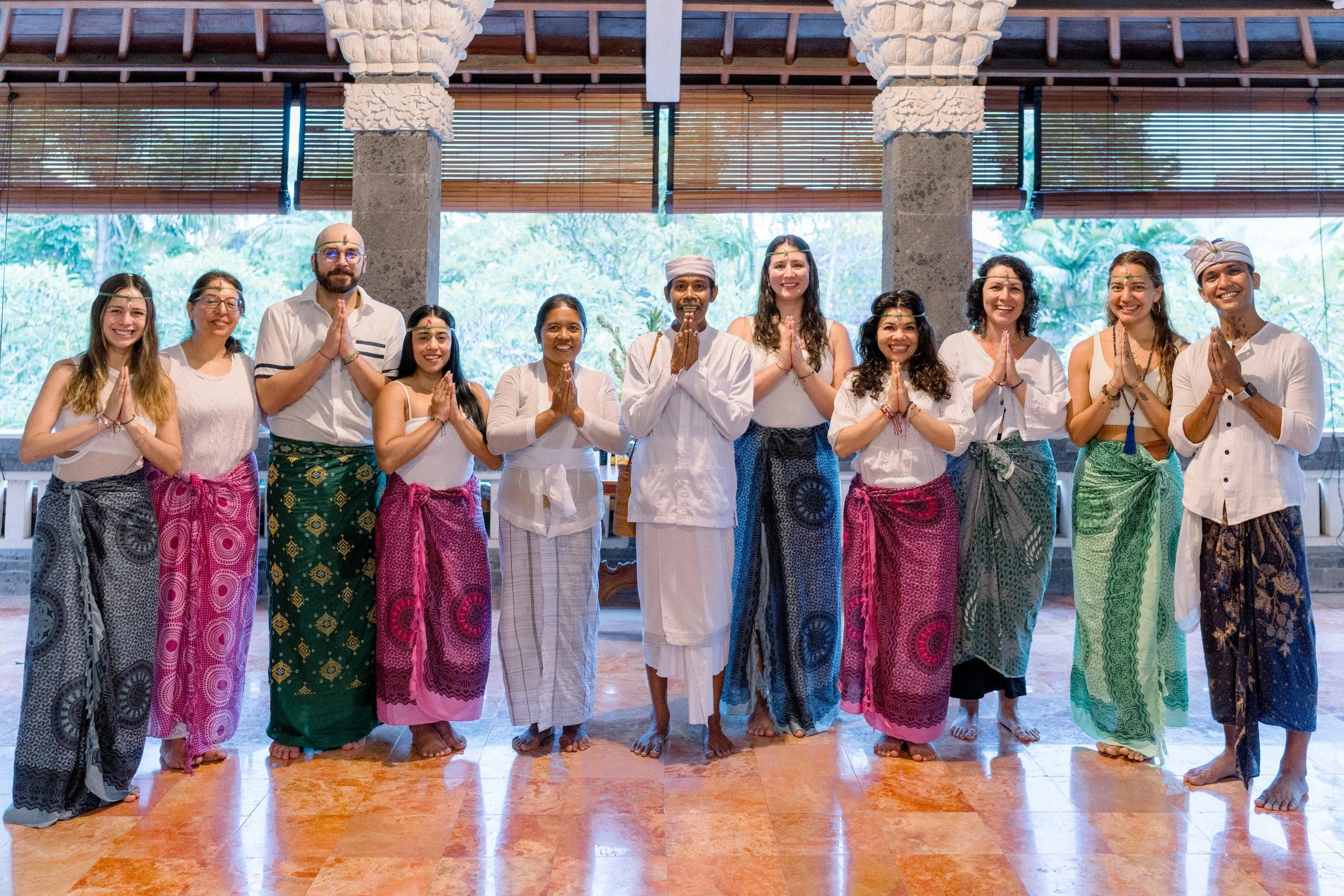 Grupo  de personas en retiro en Bali con ropa tradicional, posando con las manos juntas en señal de respeto, en un entorno interior con vista a la naturaleza.