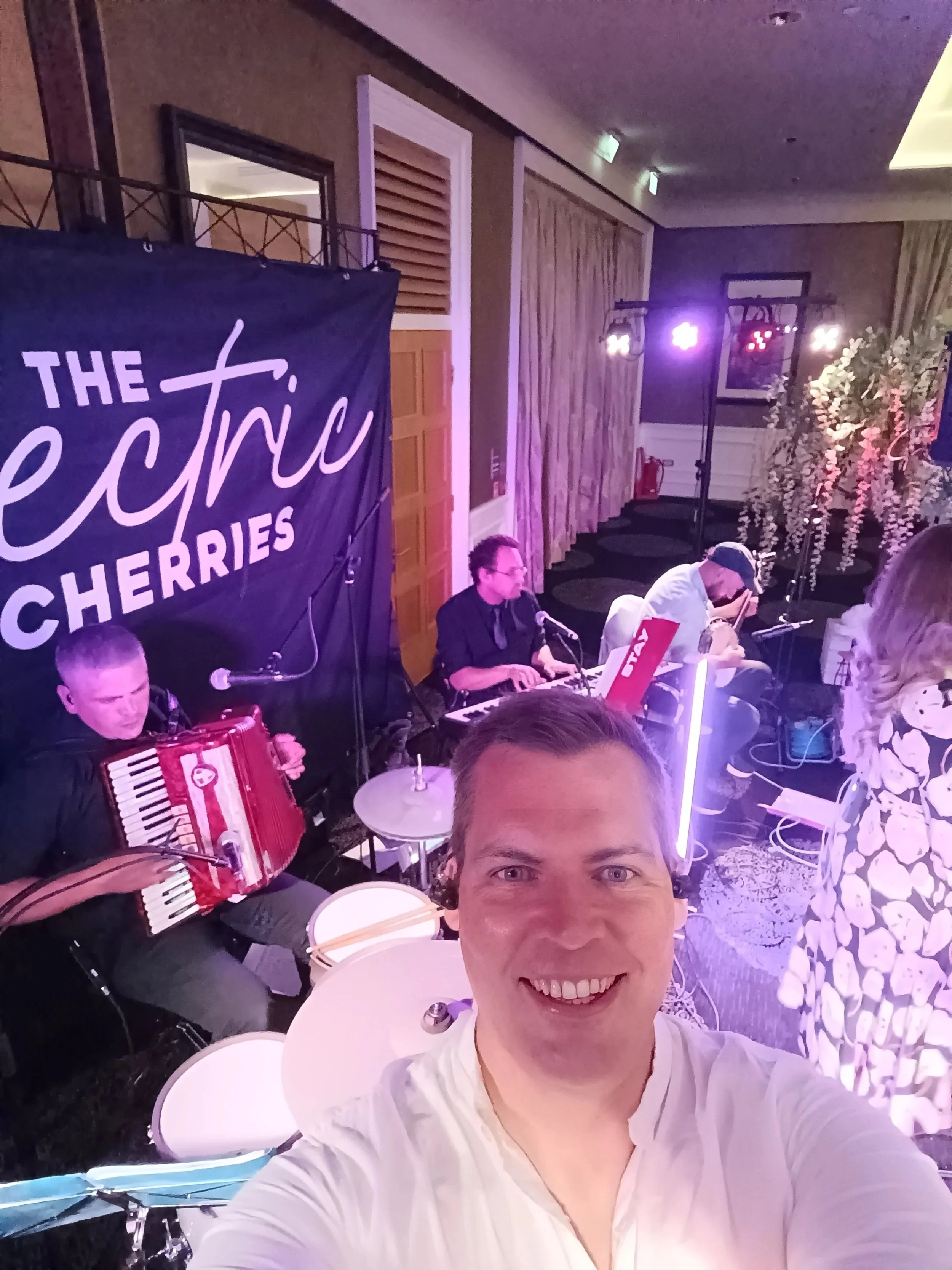 A man taking a selfie at a live music event featuring a band called 'The Electric Cherries' with members playing accordion, keyboard, and guitar in the Kingsmills Hotel Inverness. Ceilidh, wedding band, Highland, live music