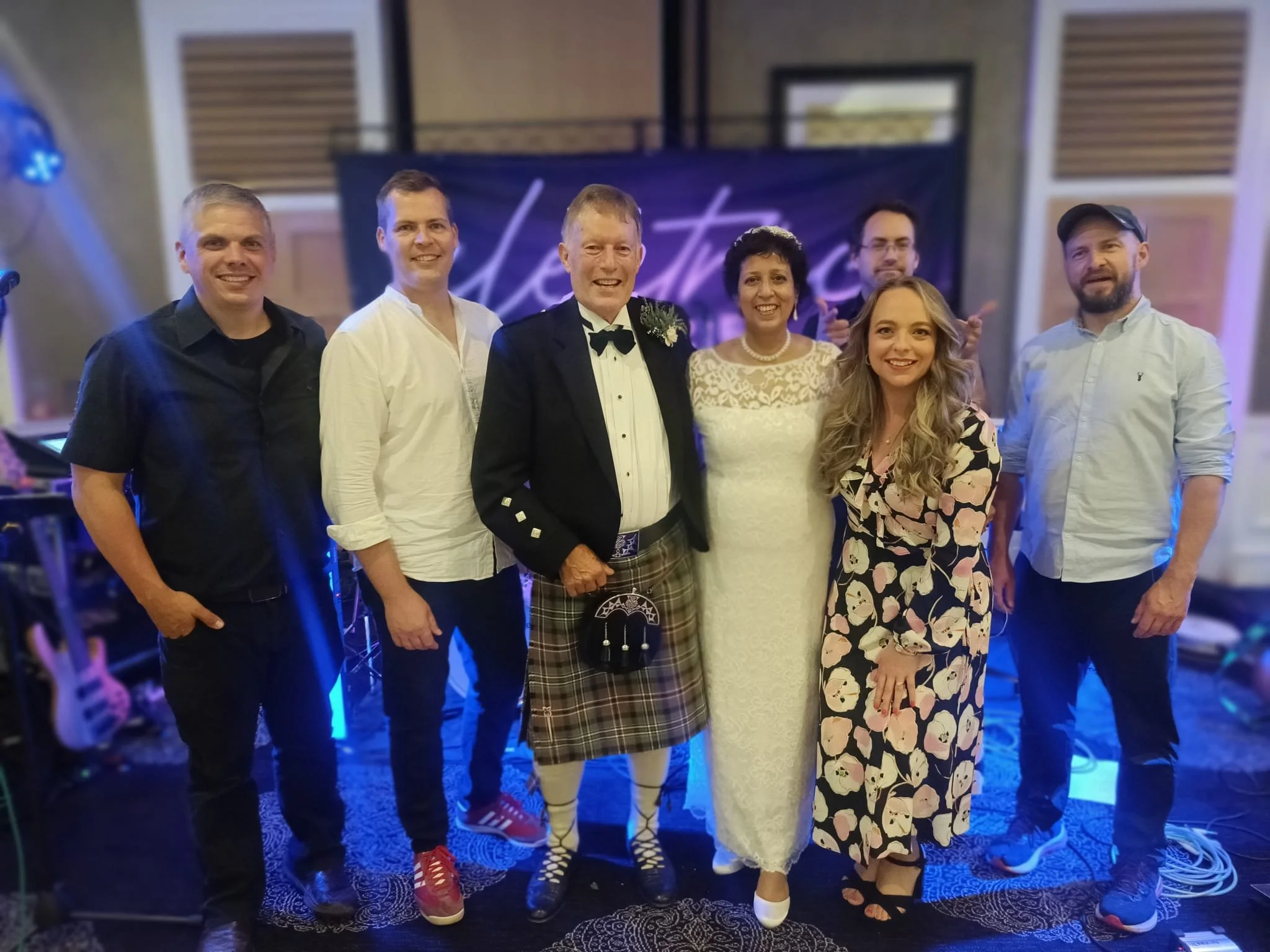Group of seven people standing on a stage, event with musical instruments visible, including guitars; curtains and a banner in the background with partially visible text.