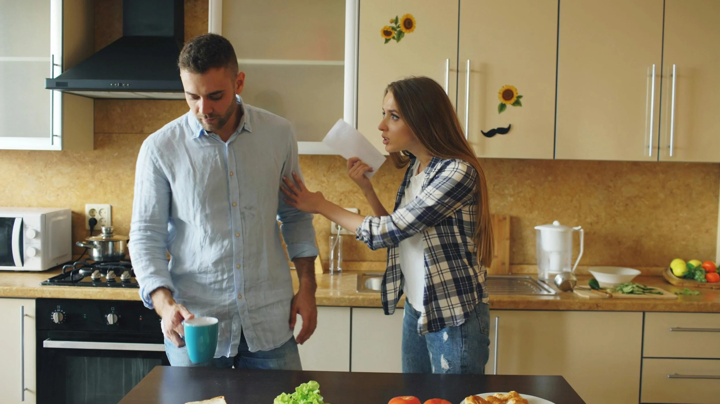 A couple in a kitchen during a conflict, with the woman reaching toward her partner as he avoids eye contact, illustrating defensiveness in relationships and marriage counseling in Worcester.