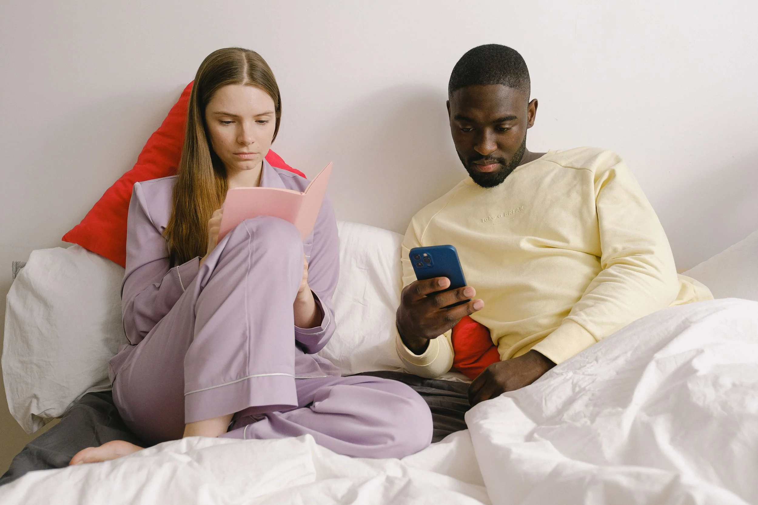 A male and female couple sitting in bed where the male is looking at his phone and the female is looking at a book.