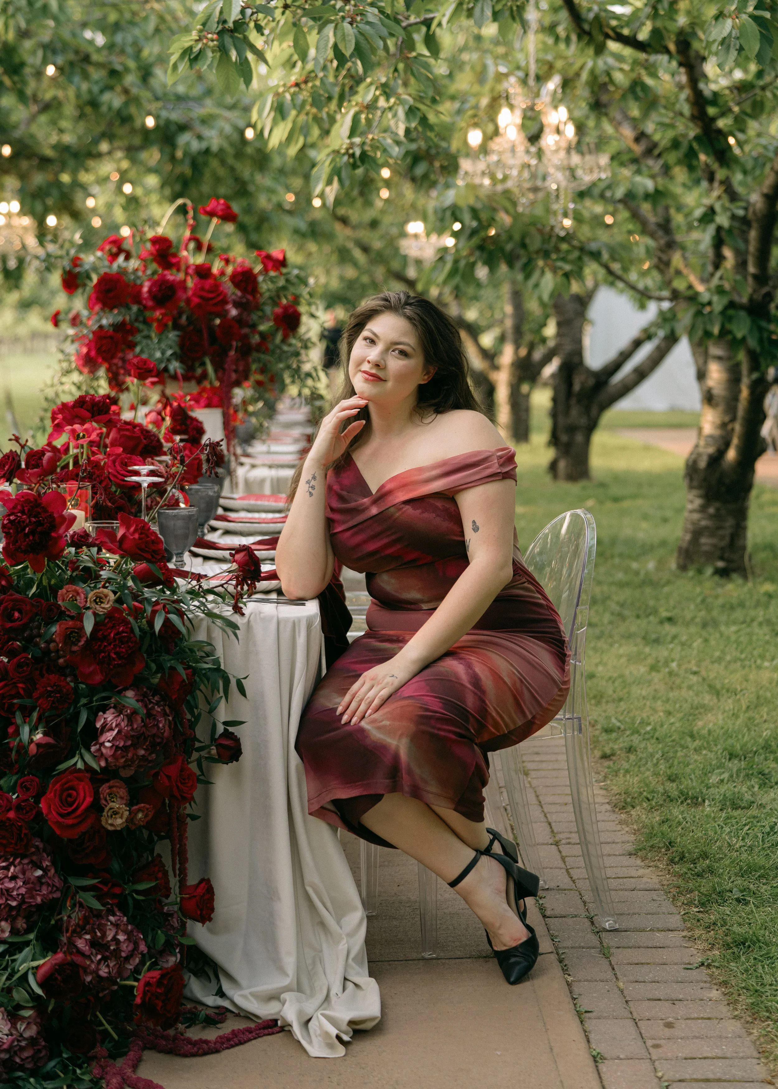 a woman dressed in a red dress sitting at a table scape featuring red dramatic florals and a white linen outdoors