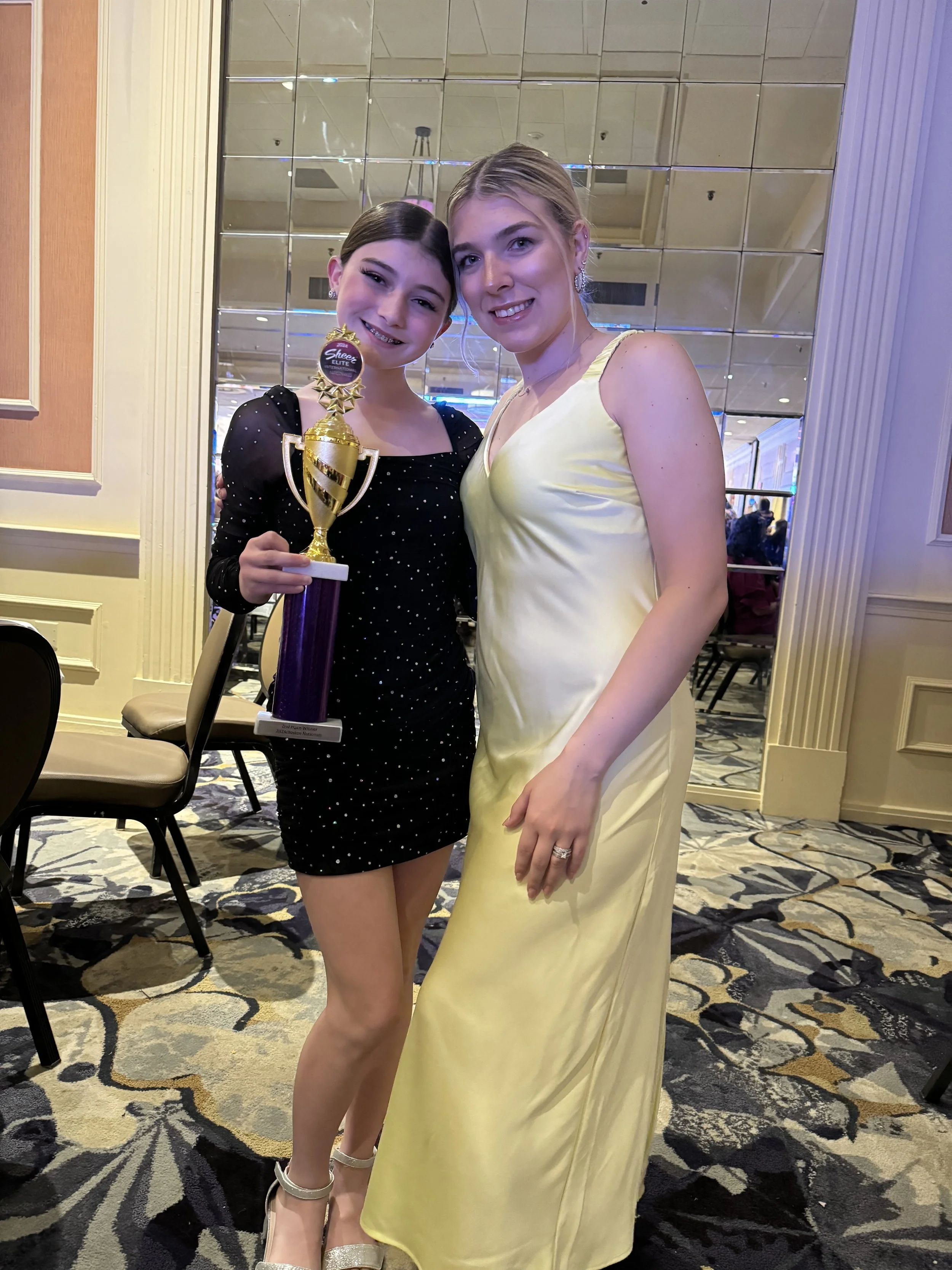 Two women standing together at an event, one holding a trophy, smiling for a photo in an elegant venue.