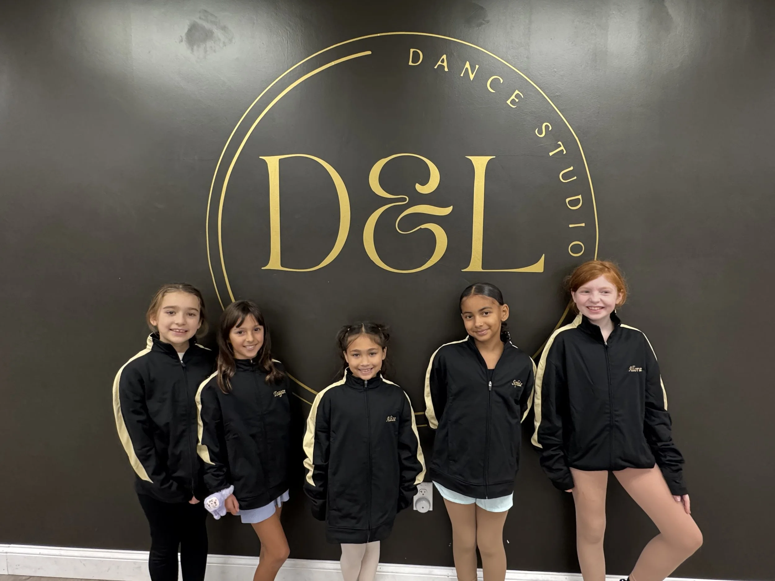 Five young girls wearing black and gold dance team jackets, standing in front of a black wall with a large gold logo reading 'D&L' and the words 'Dance Studio' in a circular design.