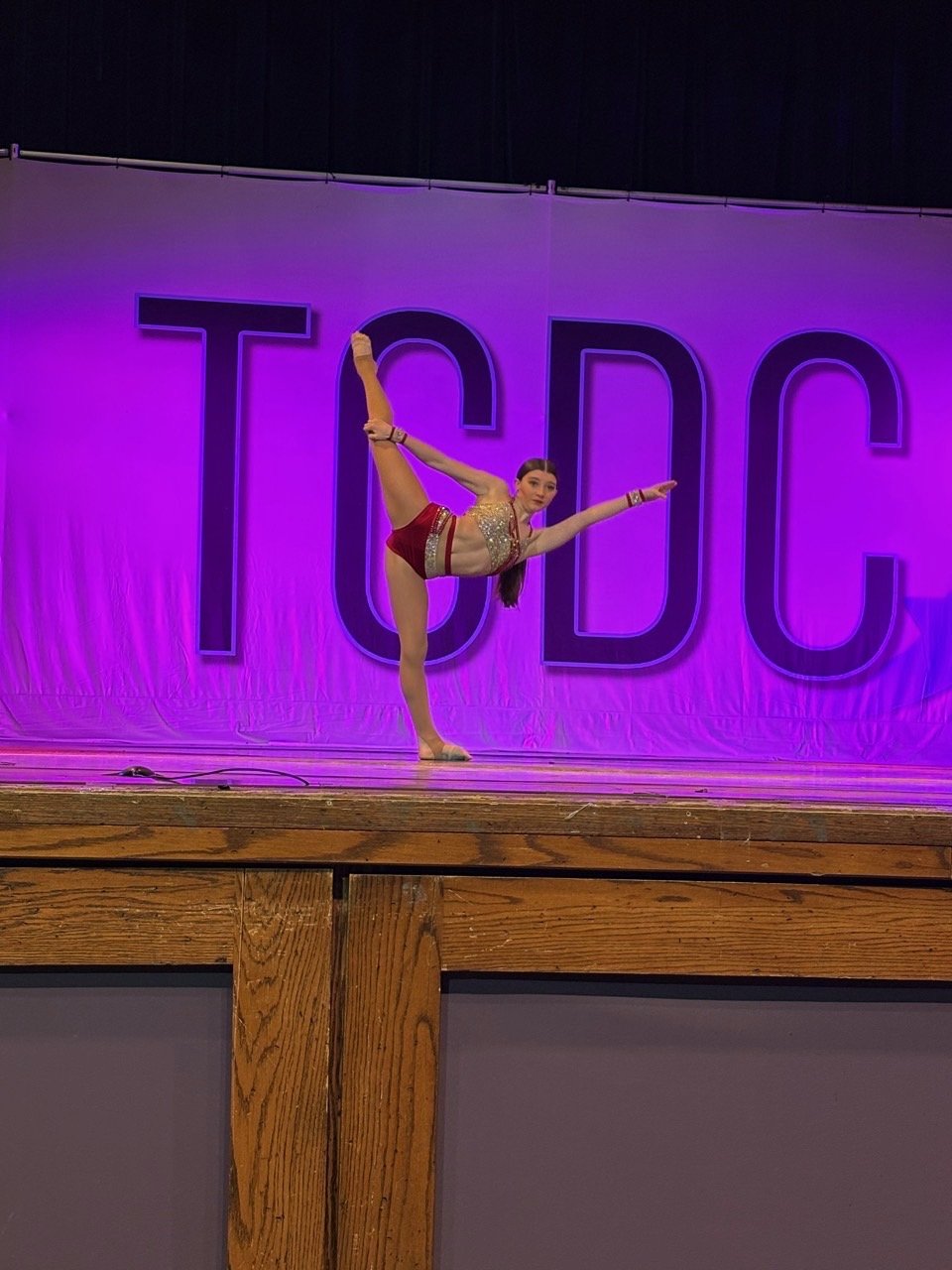 A young girl doing a gymnastics pose on stage with a large TDC sign in the background, illuminated with purple lights.