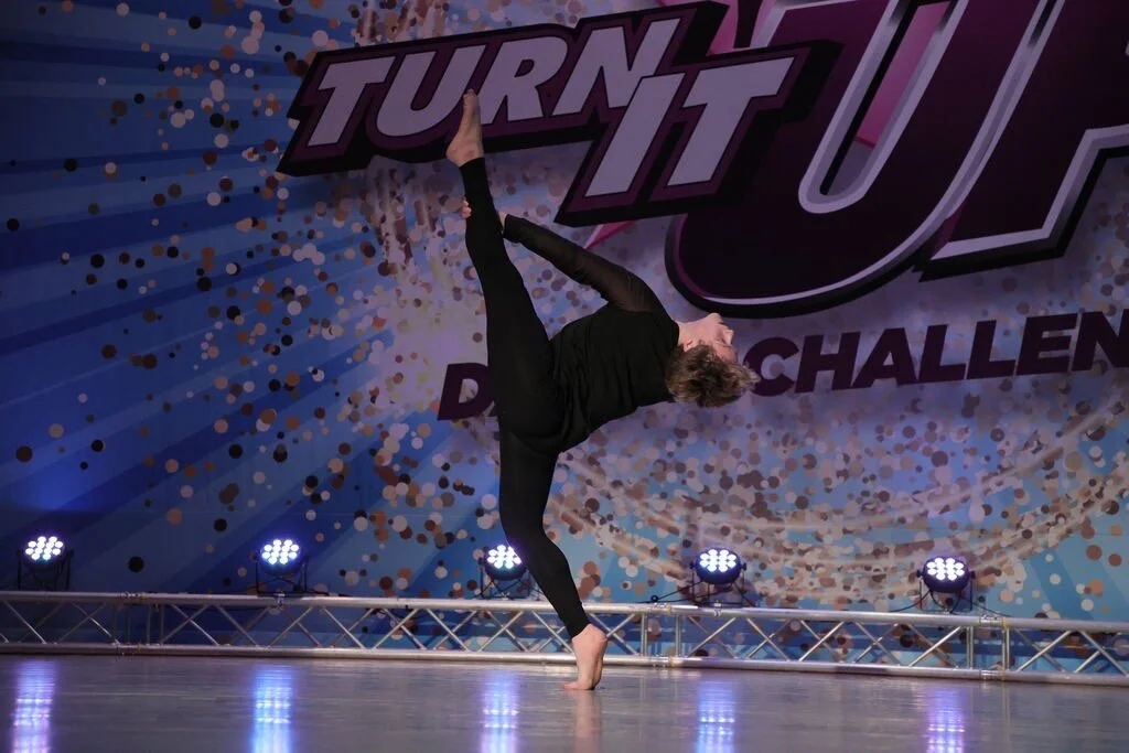 A person performing a handstand on stage during a dance or gymnastics competition, with a backdrop reading 'Turn It Up' and 'Dance Challenge', decorated with confetti and stage lights.