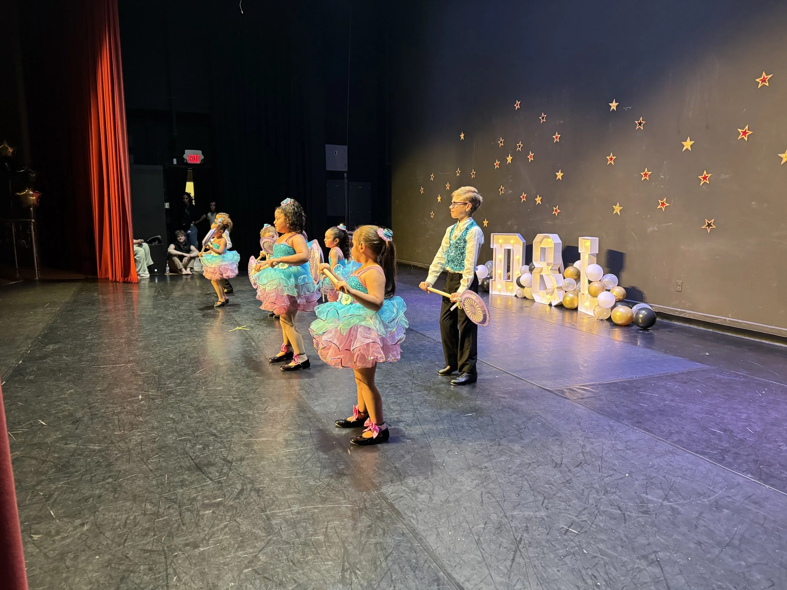 Children dressed in colorful costumes performing a dance on stage during a celebration or recital, with balloons and star decorations in the background.