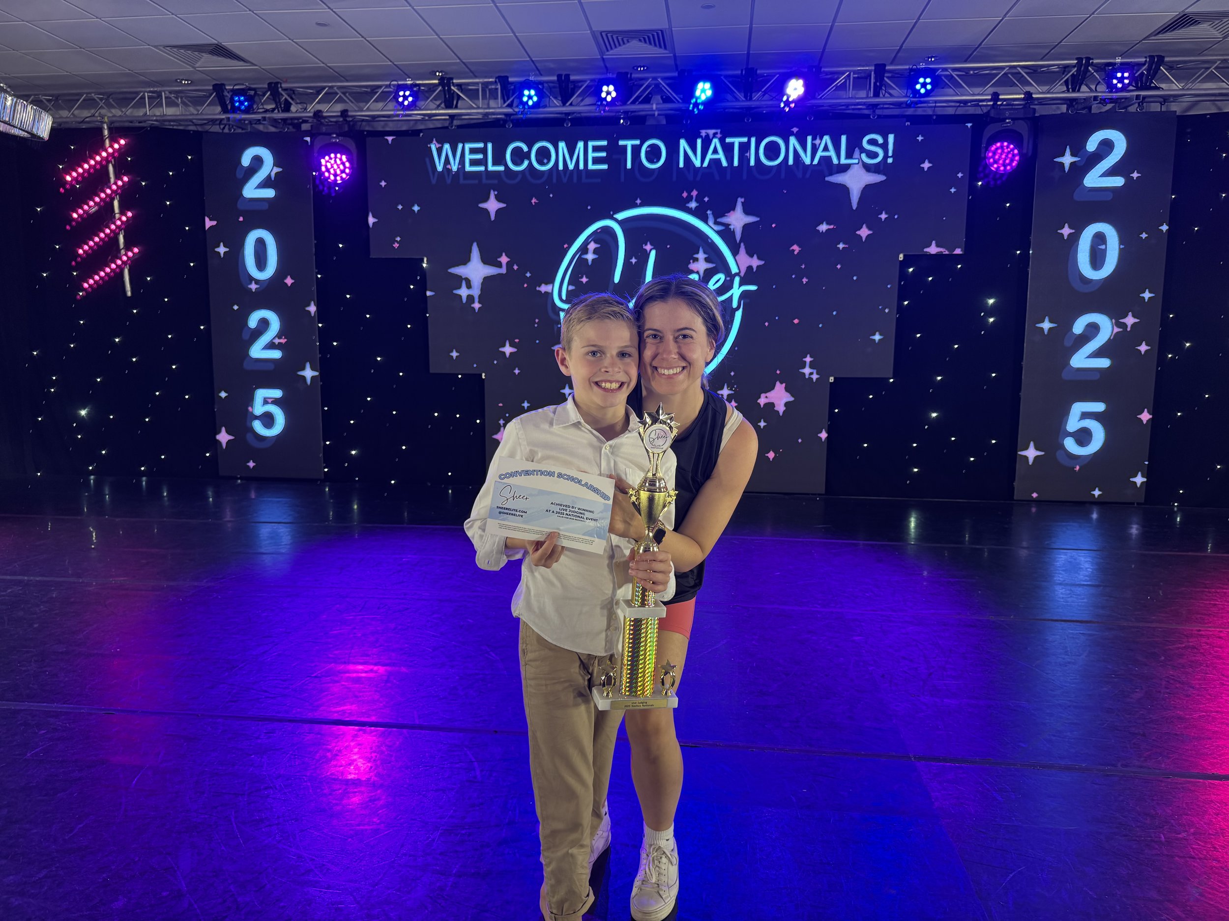 Young boy and girl holding a trophy and certificate on stage at a cheerleading competition in 2025 with a sign reading 'Welcome to Nationals'.