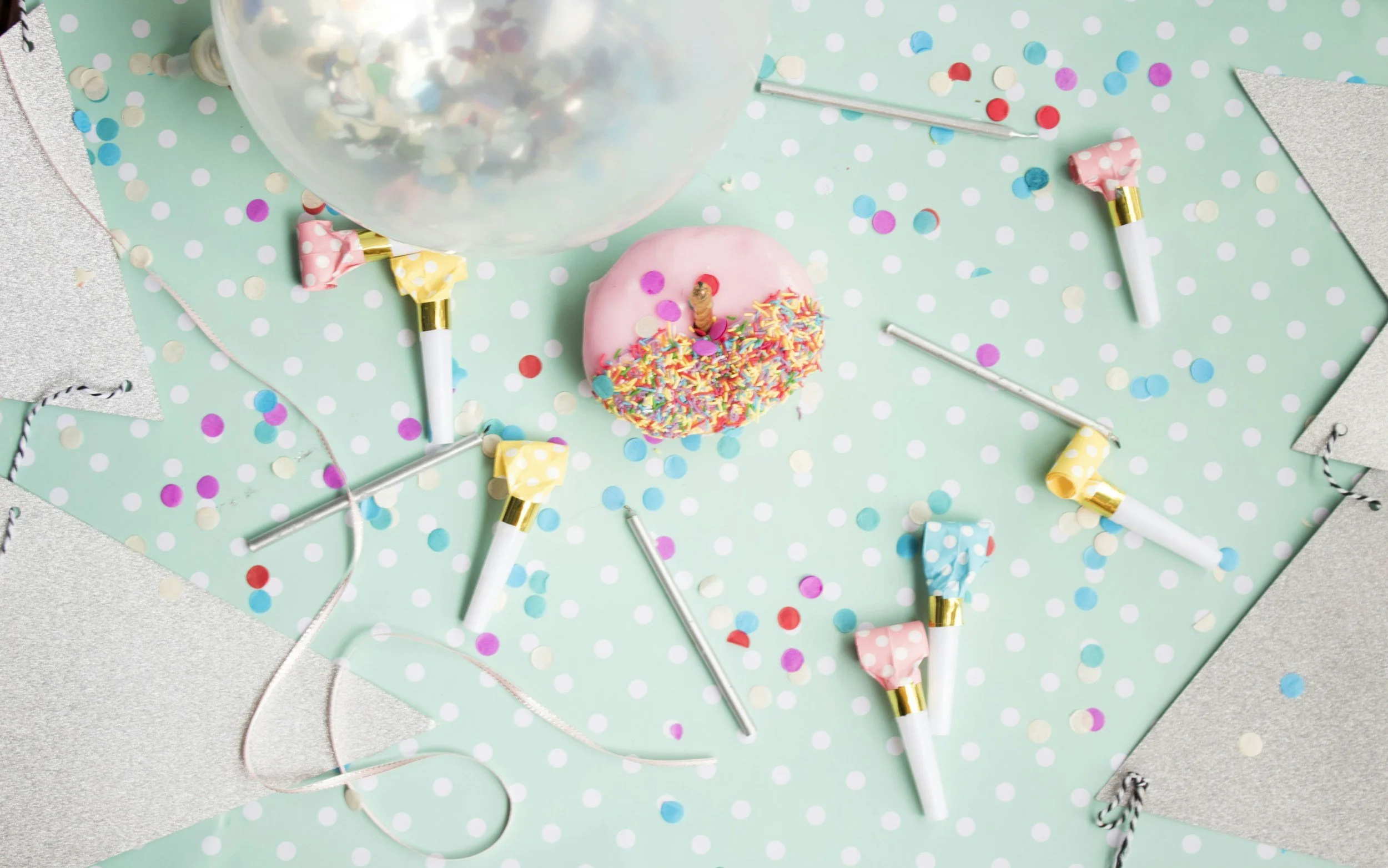 A pastel birthday party scene with a pink frosted donut with rainbow sprinkles, a clear balloon, colorful confetti, and party blowers on a light blue polka dot tablecloth.