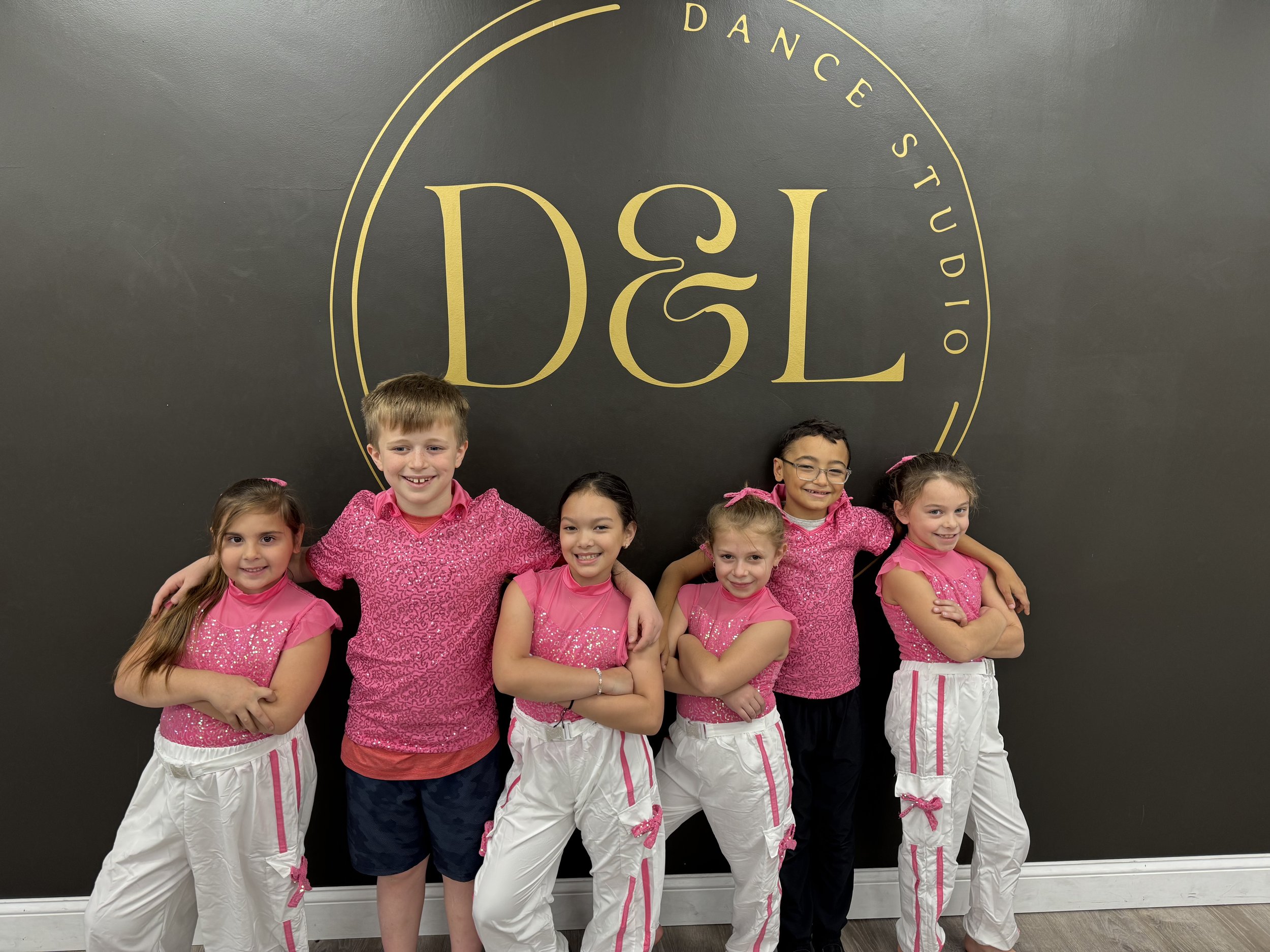 Group of six children standing in front of a black wall with a golden logo reading 'D&L Dance Studio.' The children are dressed in pink and white dance costumes, with most smiling and some with arms around each other.