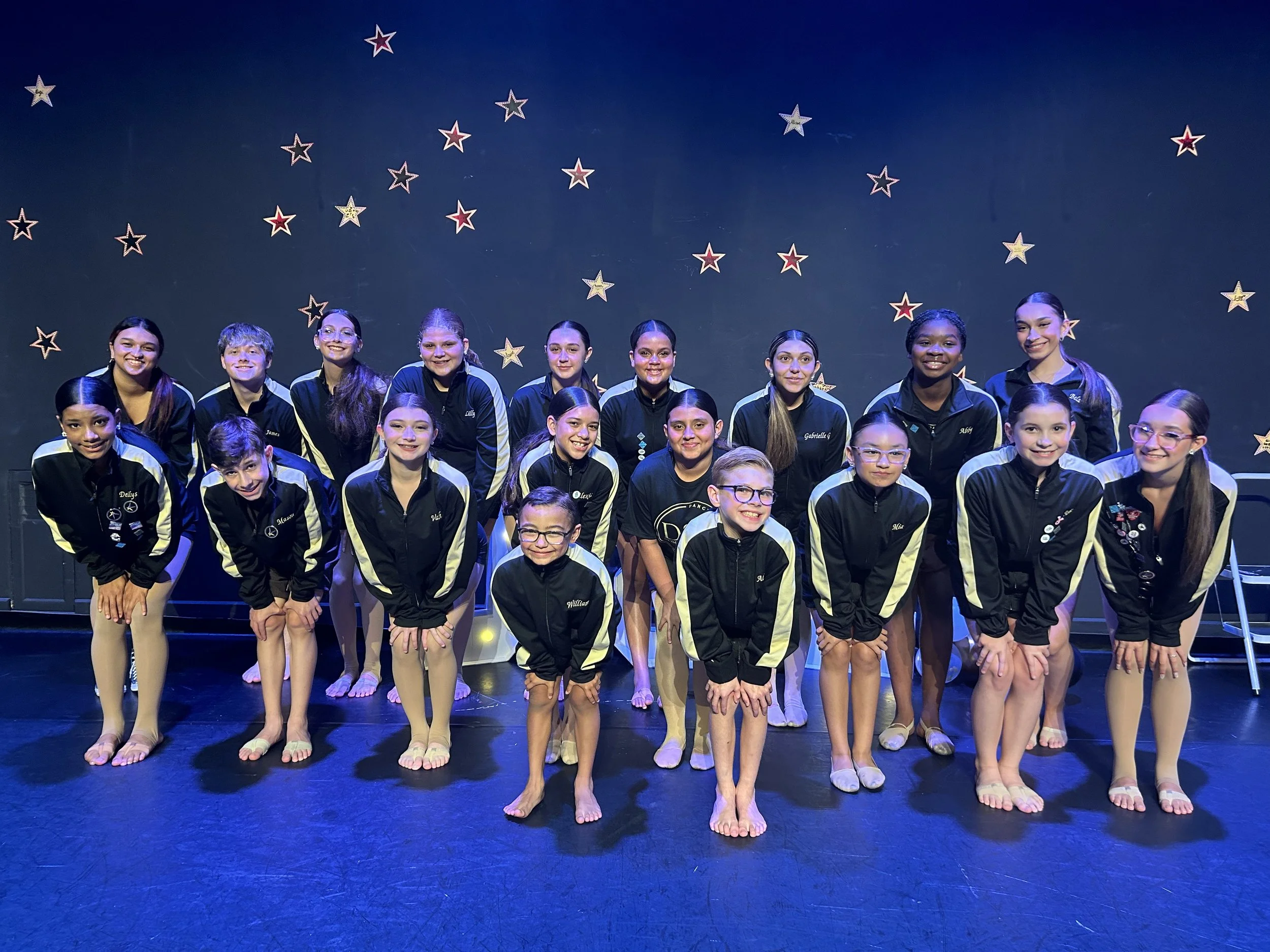 Group of young girls dressed in black and gold jackets, posing on stage with star decorations on the black backdrop.
