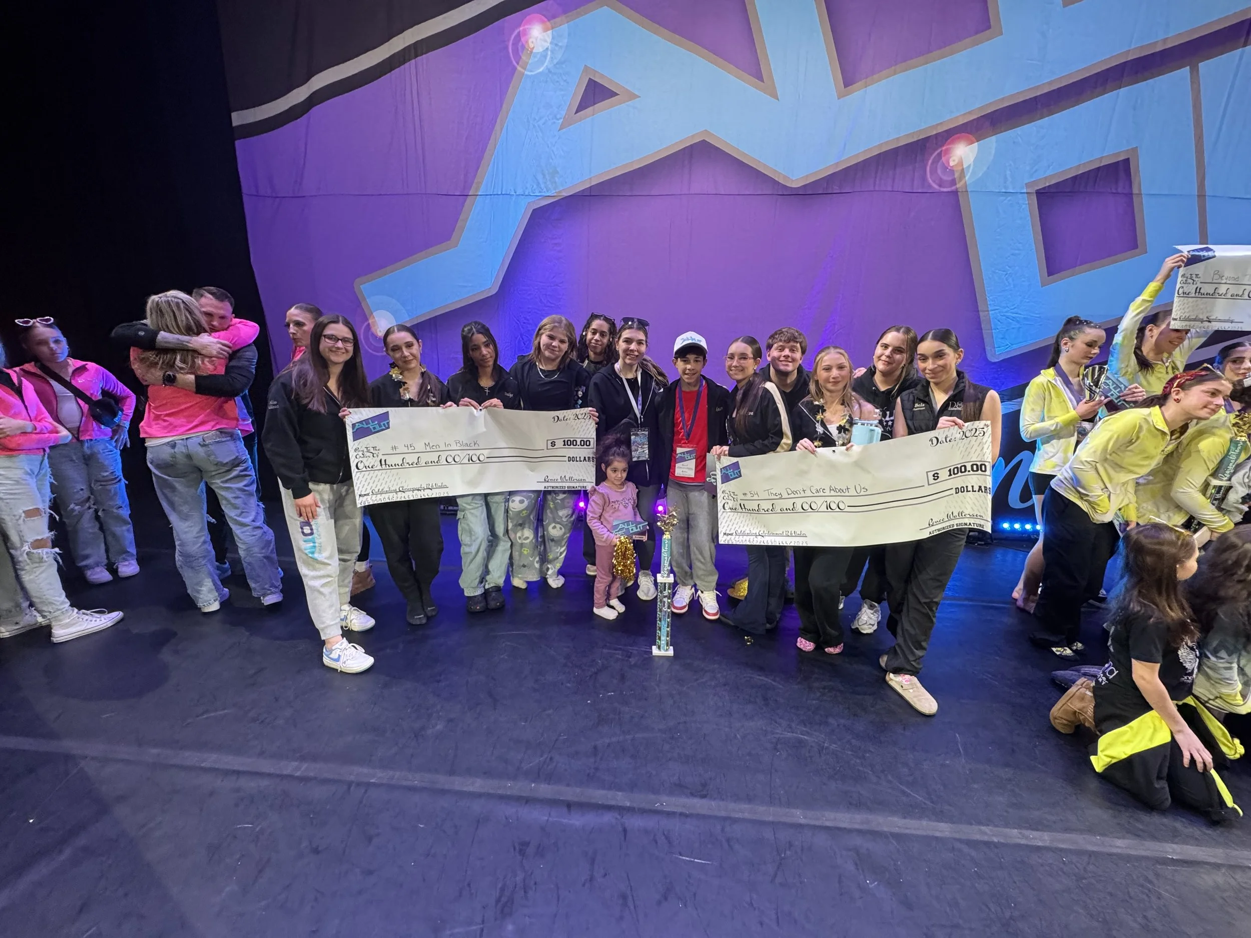 A group of young performers on a stage holding large checks and smiling, celebrating a victory, with a large colorful backdrop behind them.
