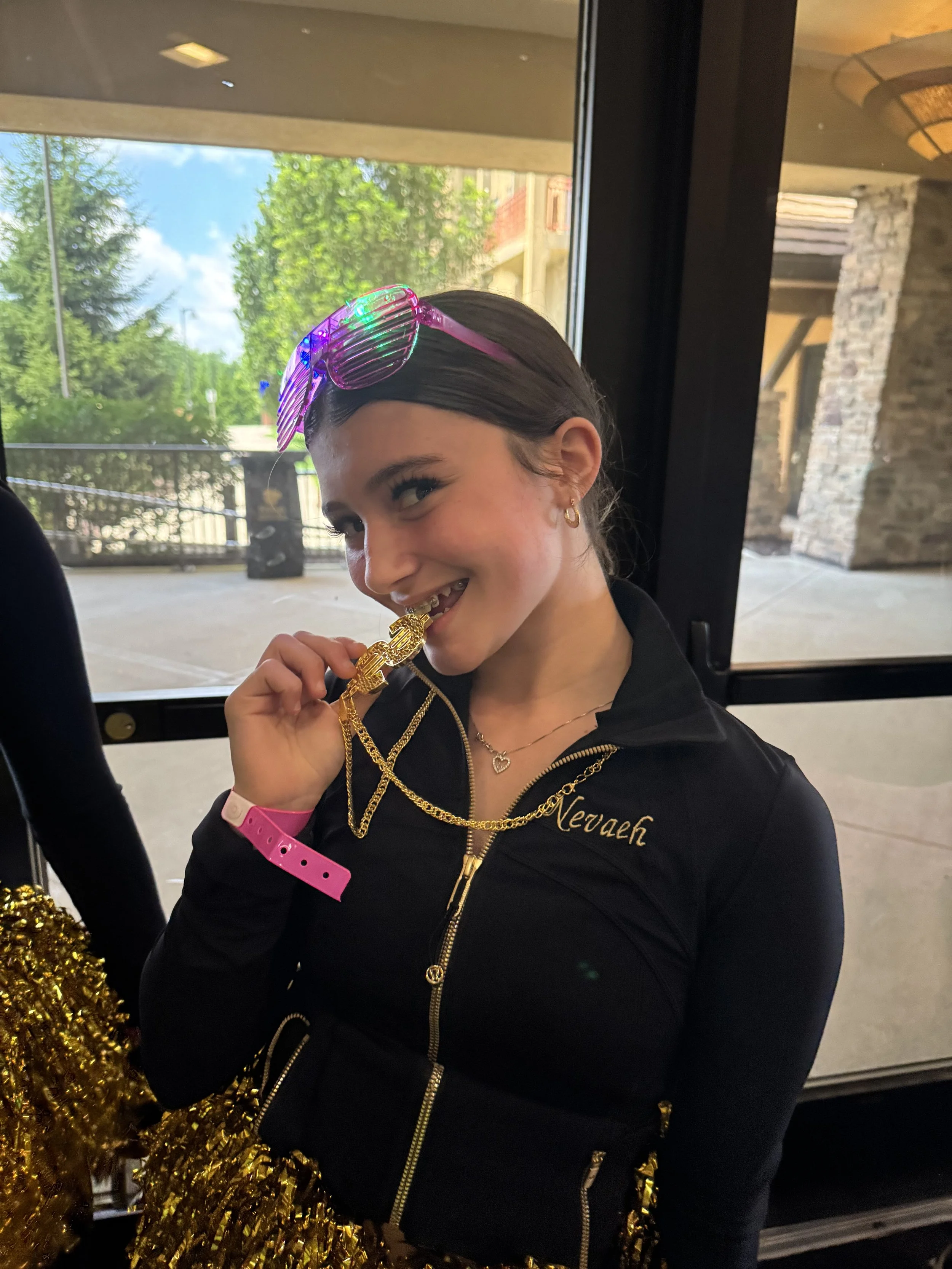A young woman with dark hair, wearing rainbow-colored sunglasses on her head, a black jacket with gold zippers and a gold embroidered name, gold jewelry, and a bright pink wristband, is smiling and biting a gold chain necklace inside a building near 