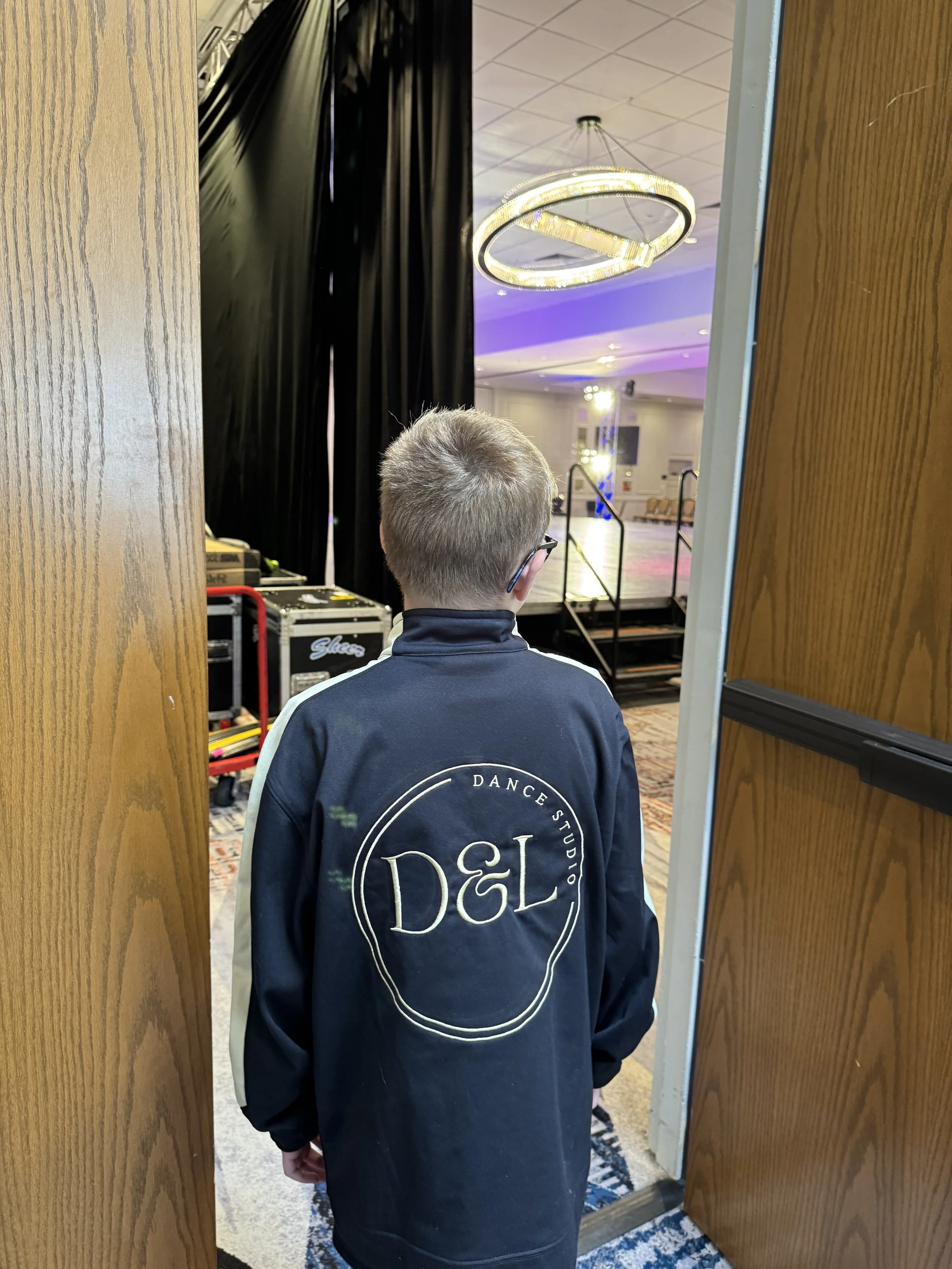 Back of a young person with short blonde hair wearing a black jacket with 'DANCE STUDIO &L' on the back, standing in a doorway looking toward a staged event area with black curtains, rolling equipment cases, and overhead lighting.