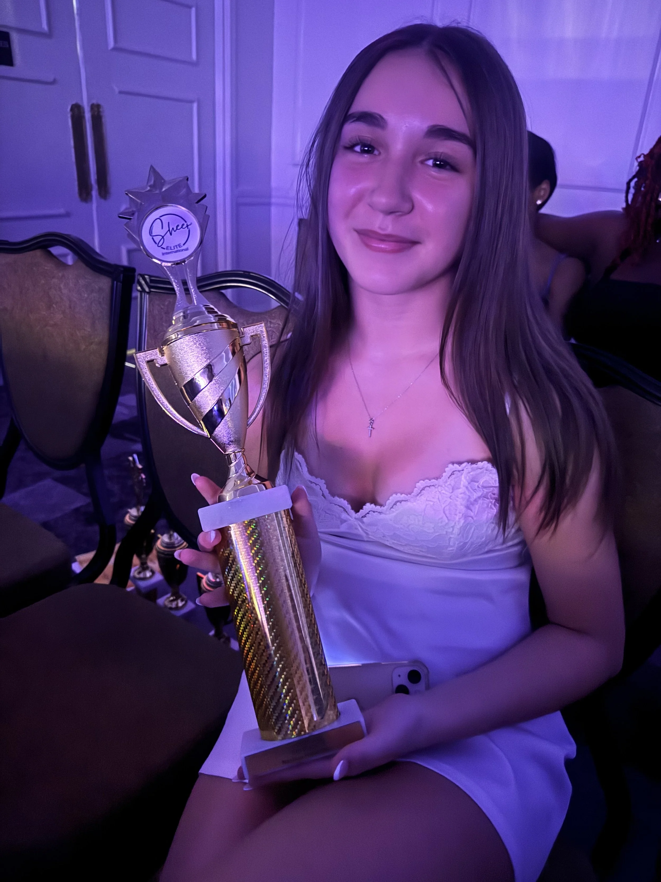 Young woman in white dress smiling and holding a gold trophy with a purple and gold ribbon, sitting on a chair in a dimly lit room.