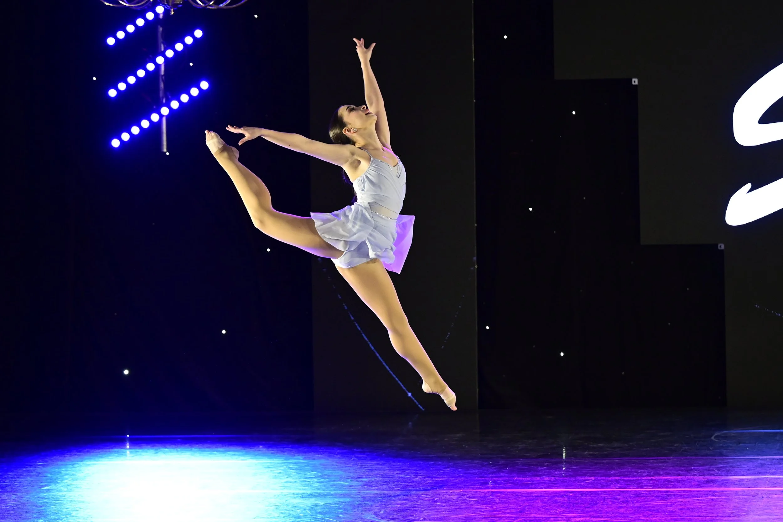 A female ballet dancer performing a jump on stage with a black backdrop and purple and blue lighting.