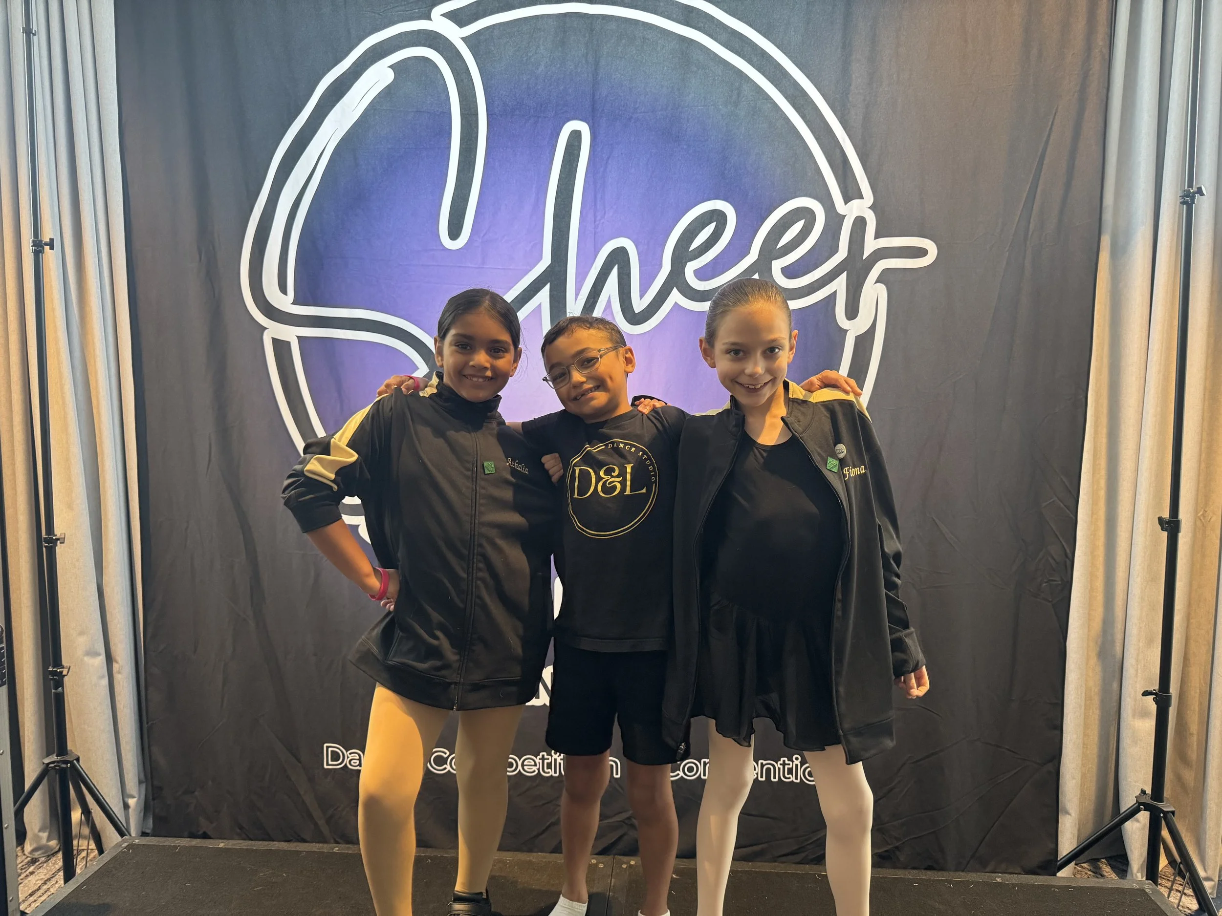 Three young girls standing side by side, smiling, in front of a black backdrop with a large white and purple logo that reads 'Shee'. They are dressed in black jackets and light-colored tights, with arms around each other.