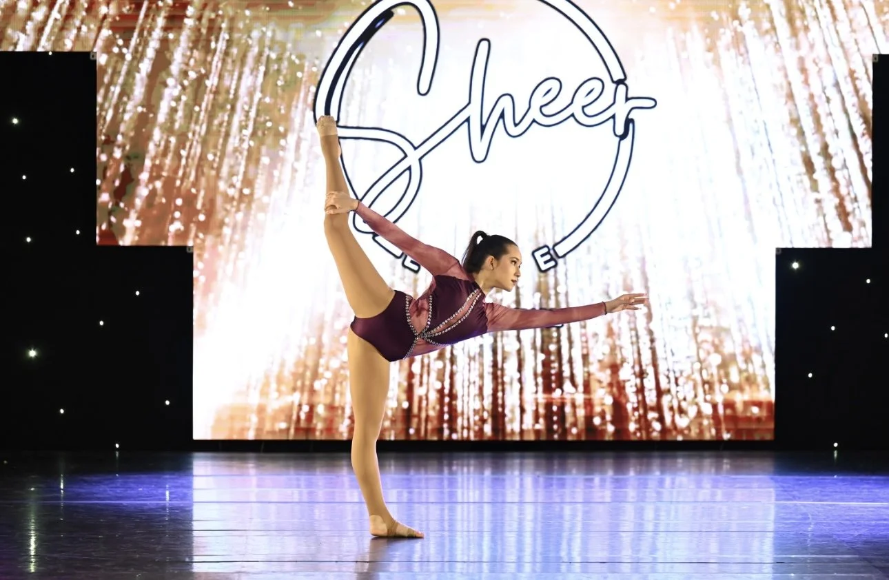 A female gymnast performing a balance pose on stage with a competition background reading 'Cheer' and decorative lighting.