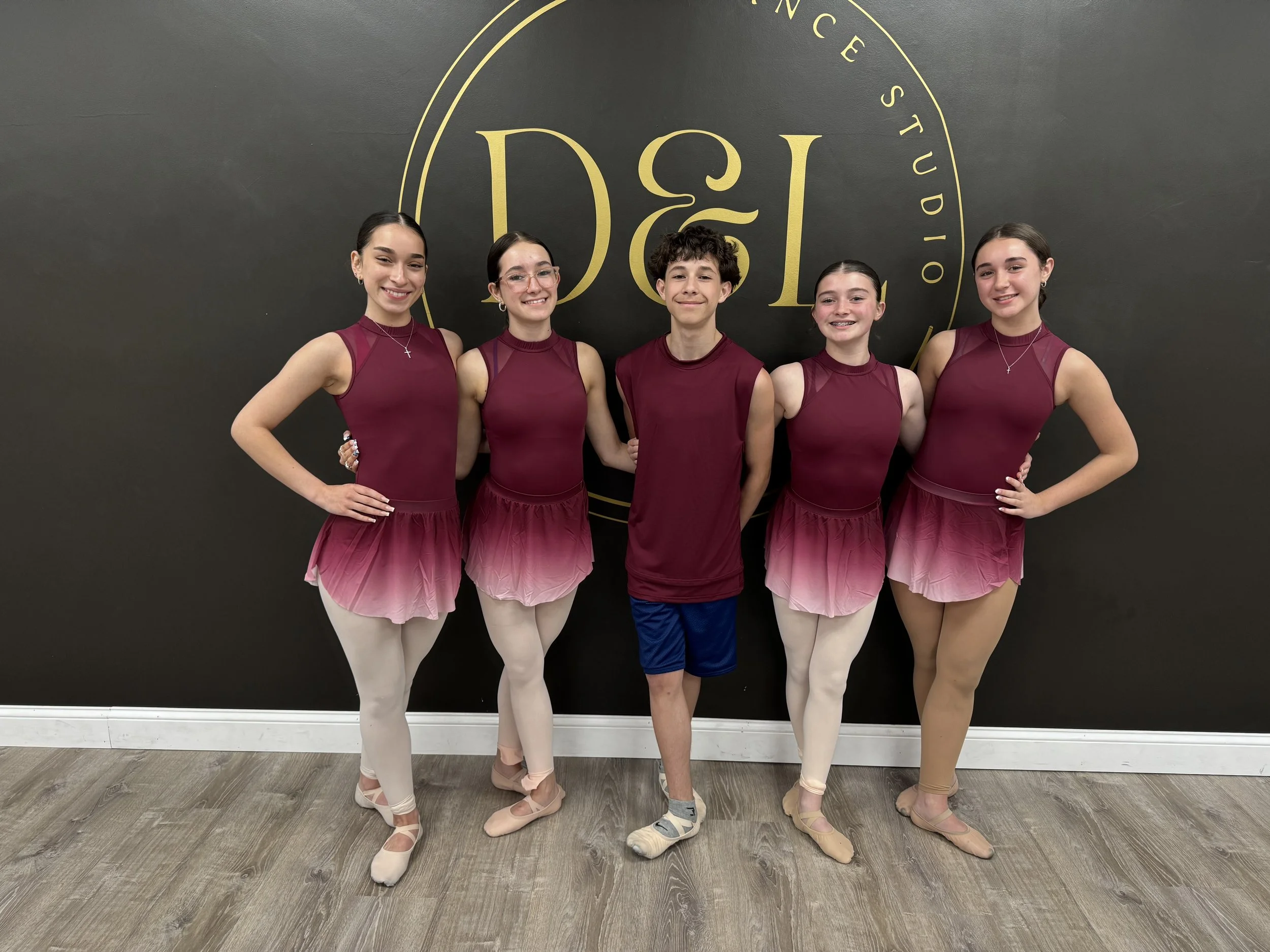 Five young ballet dancers posing together in front of a black wall with a gold logo. Four girls are wearing matching maroon and pink gradient tutu costumes and beige tights, and a boy is in a maroon tank top and navy blue shorts.
