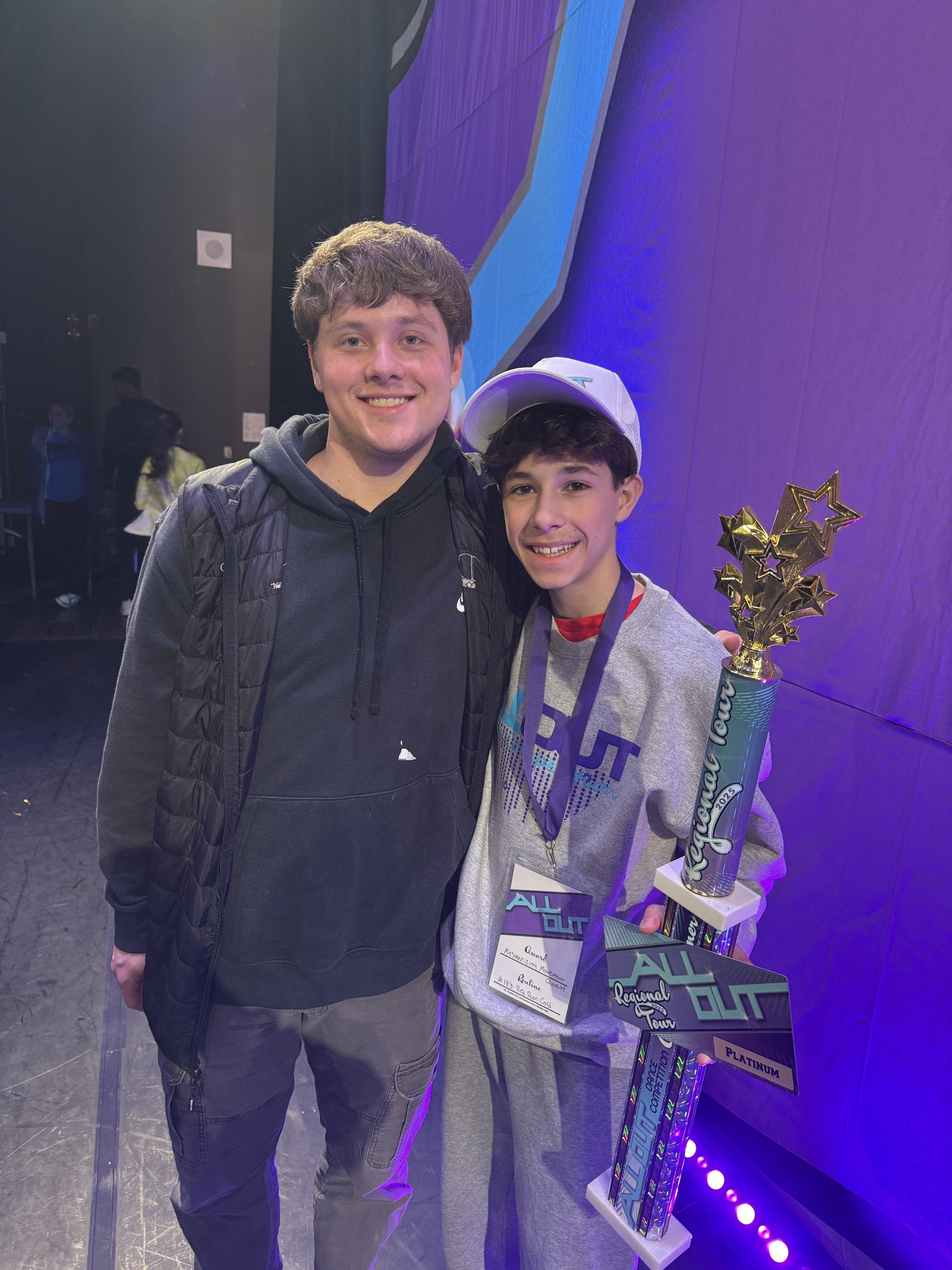 Two boys standing together, smiling, with one holding a large trophy. The boy on the right wears a gray sweatshirt, a white baseball cap, and a medal around his neck. The background has dark walls with colorful lighting.