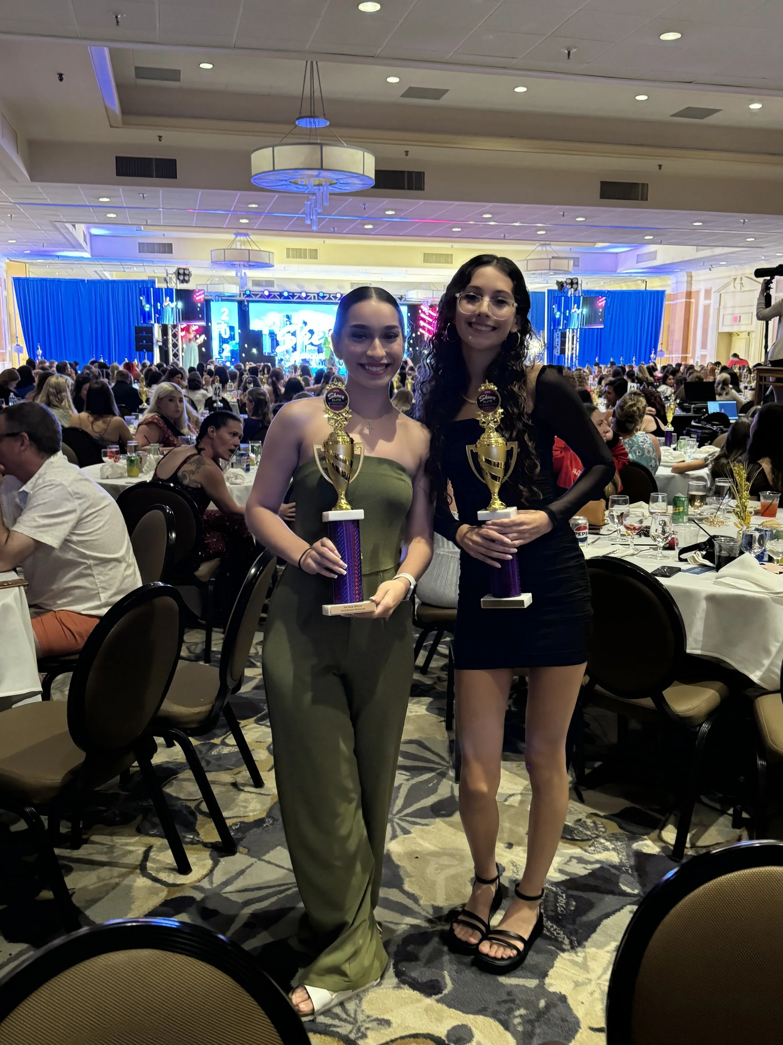 Two young women standing and holding trophies at an awards ceremony in a large banquet hall filled with seated guests.