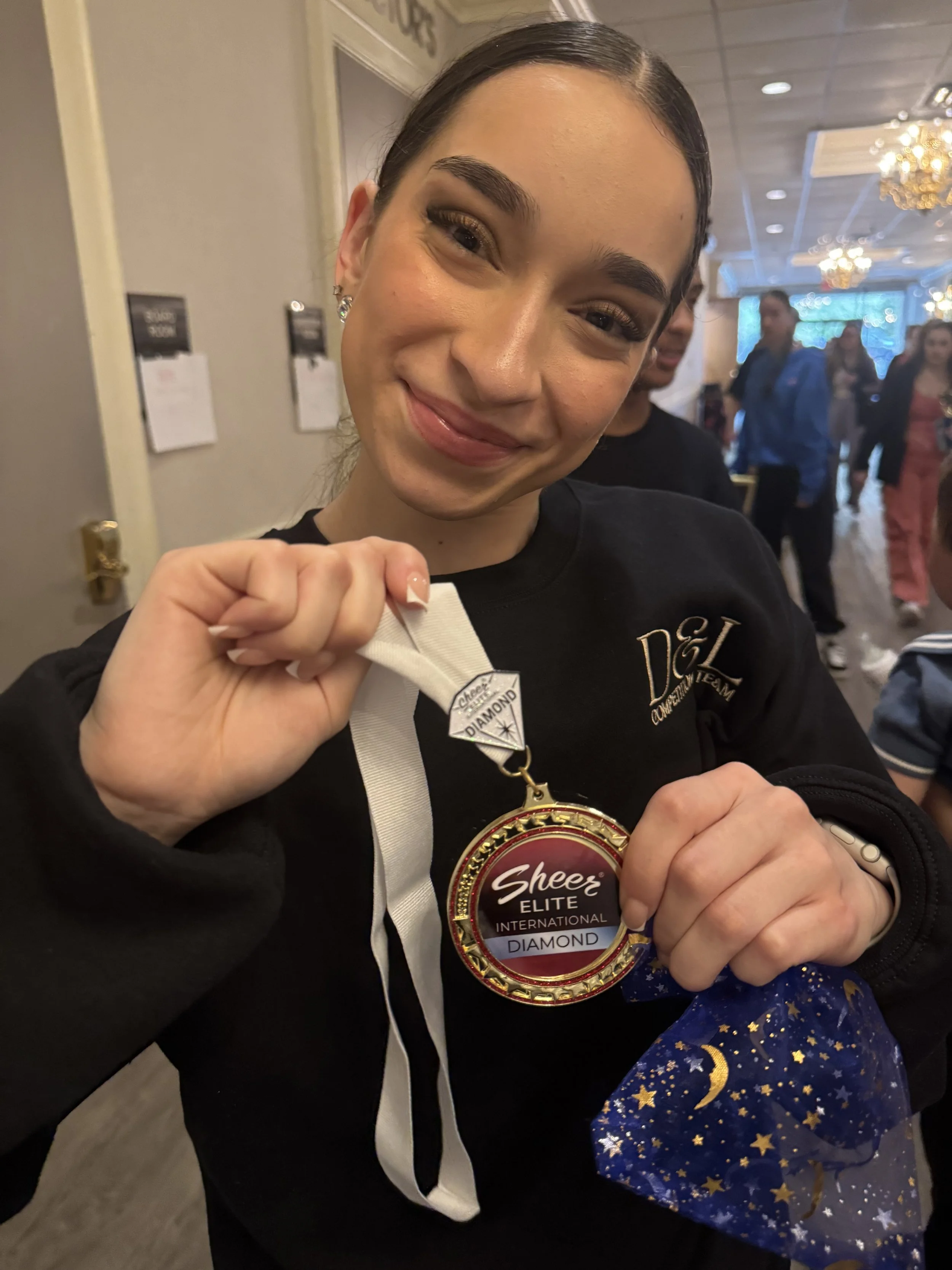 A woman smiling and holding a medal that reads 'Sheer Elite International Diamond'. She is in an indoor event space with people in the background, and there is a blue gift bag with gold stars in her hand.