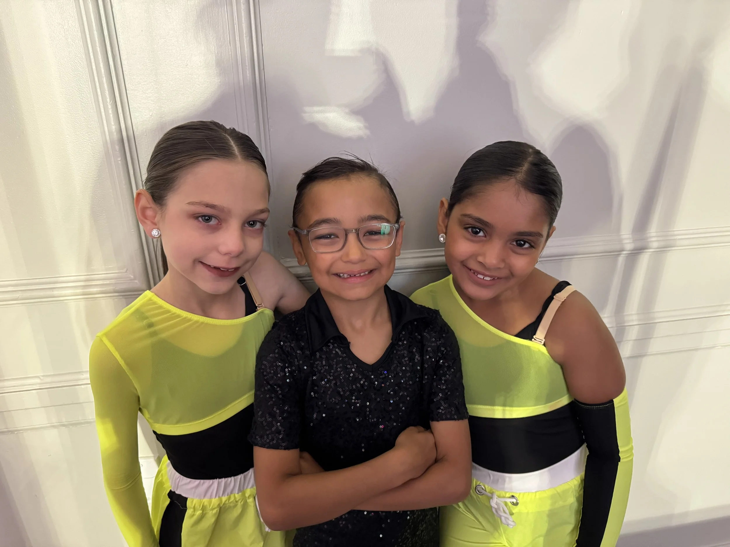 Three young girls smiling, standing side by side against a white wall. Two are wearing bright yellow and black dance costumes, and the girl in the middle is wearing glasses and a black sparkly top.