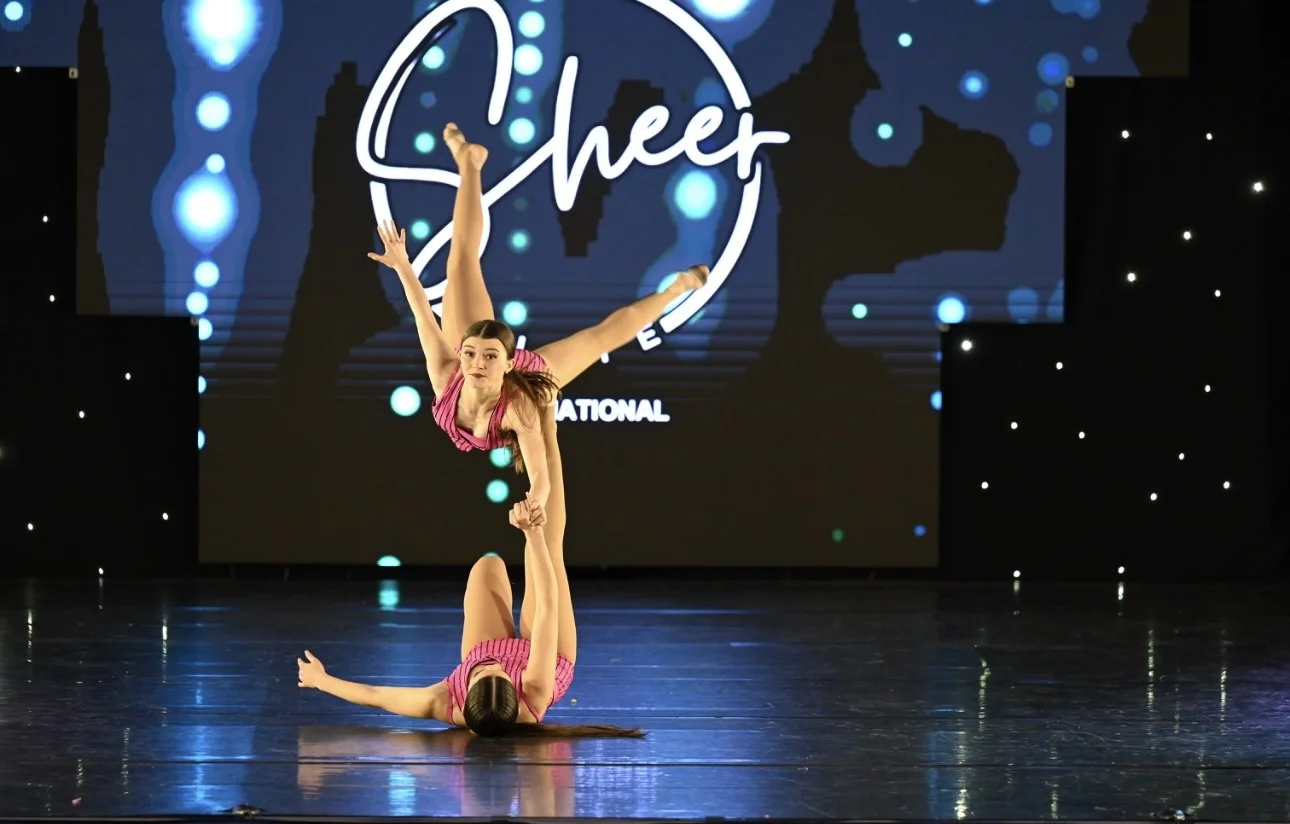 Two young girls performing an acrobatic dance routine on stage, one standing on the other's shoulders, with a blue and black backdrop featuring the Cheer brand logo.