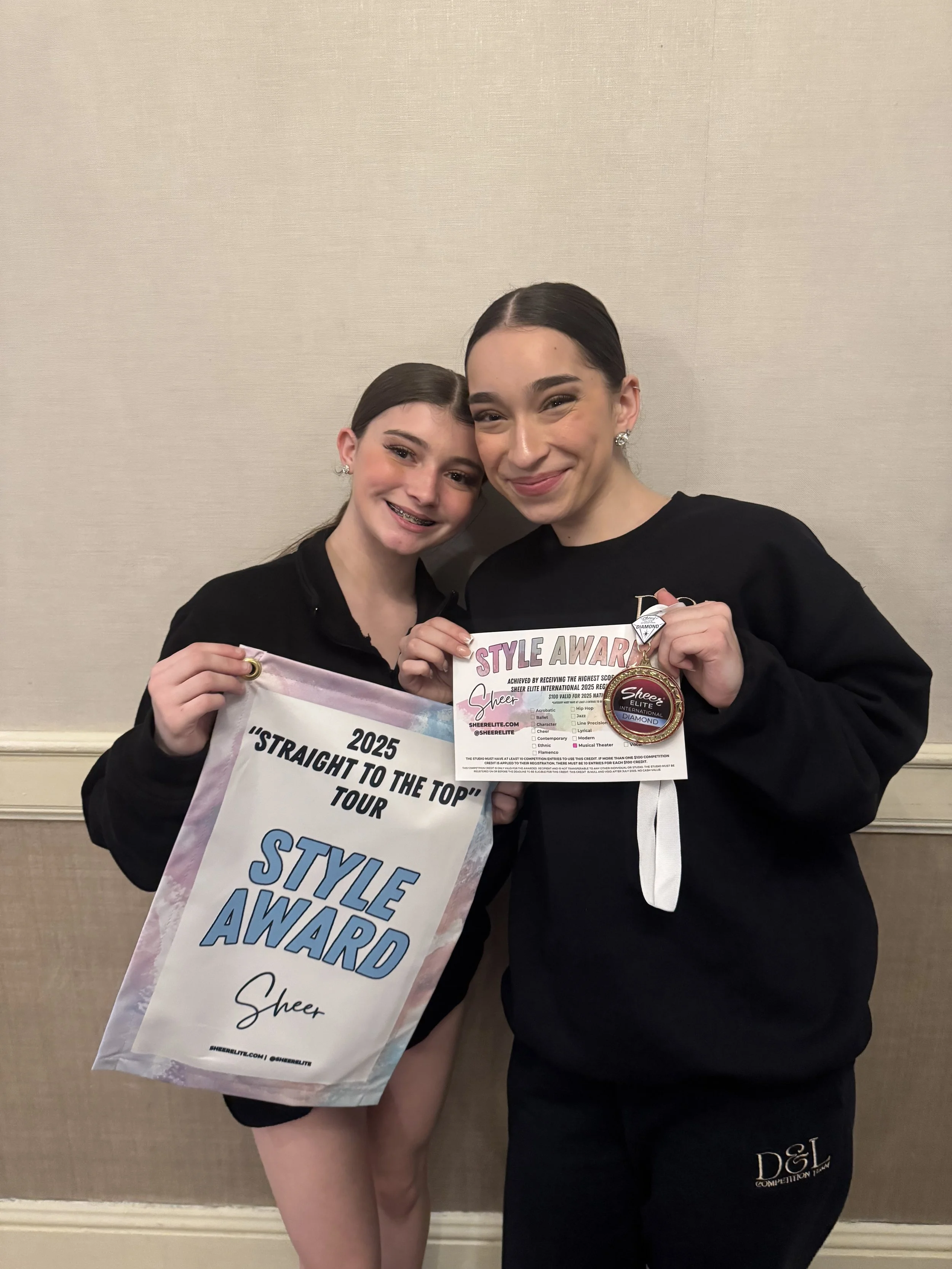 Two young female dancers smiling while holding awards at a dance competition, standing against a plain beige wall.