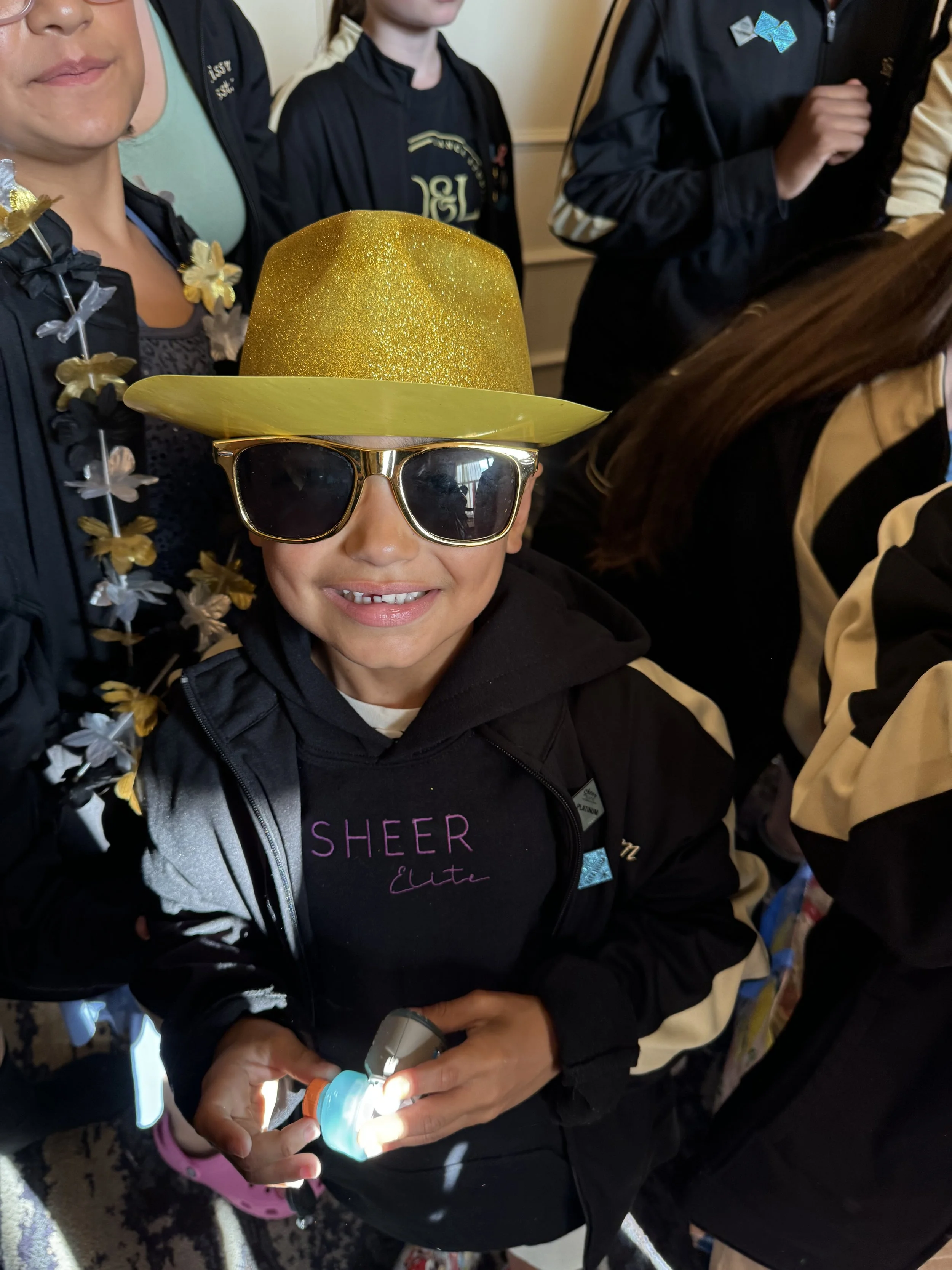 Young boy wearing a gold glittery hat, sunglasses, and a black jacket with a floral lei, holding a flashlight, surrounded by other children in black and beige jackets in a classroom or indoor setting.