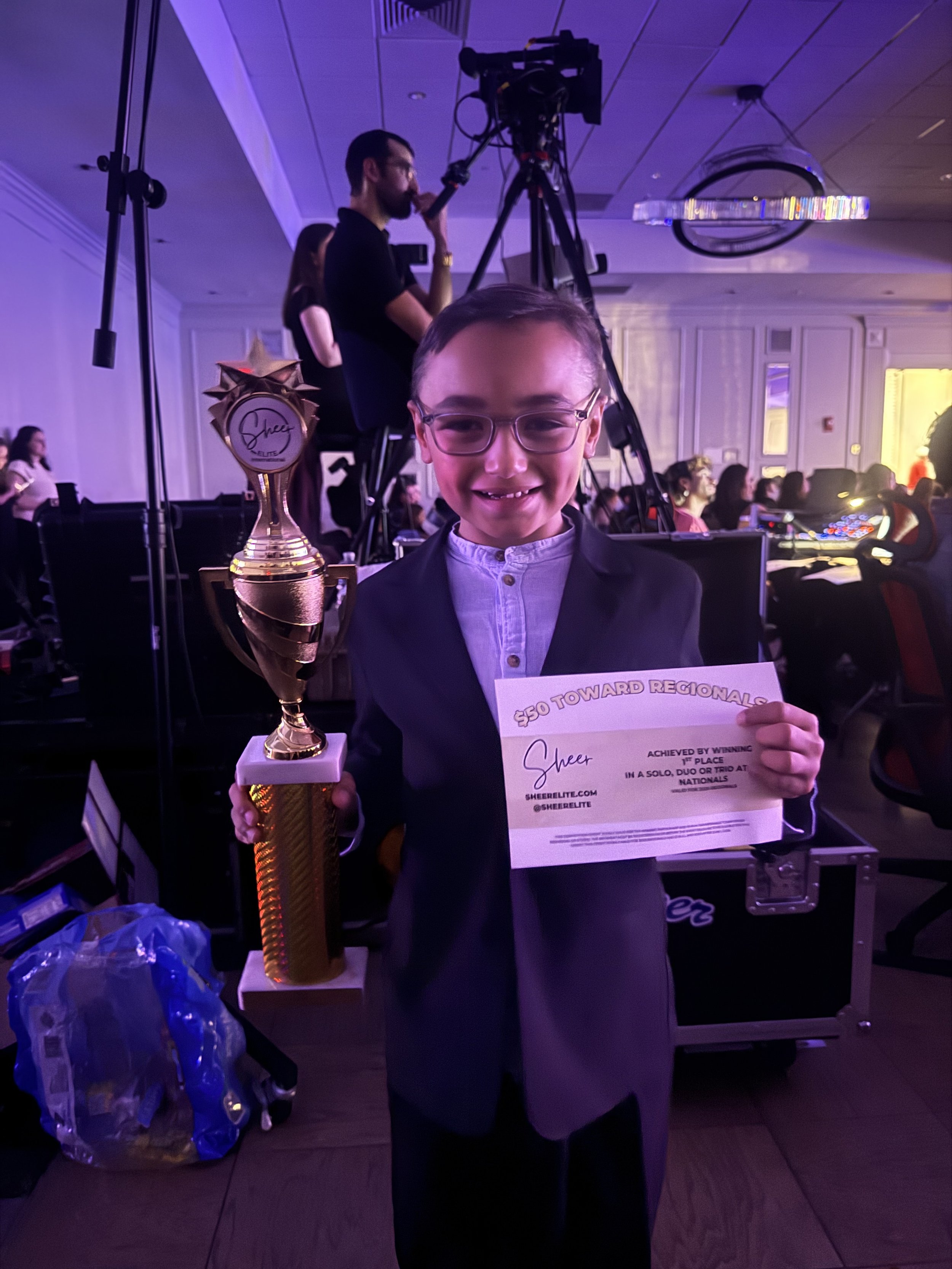 Young boy in glasses and suit holding a trophy and certificate at an awards event.