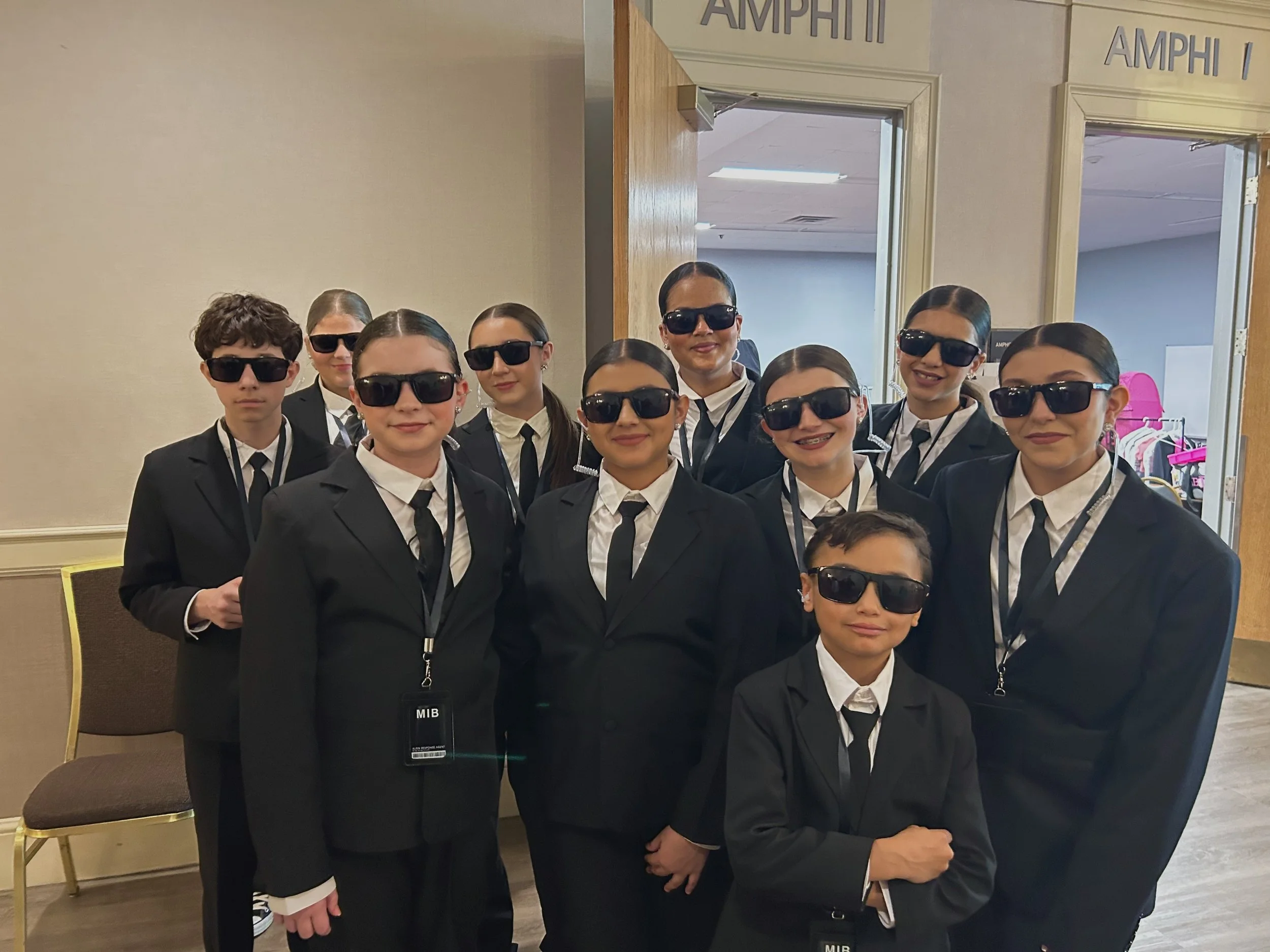 A group of children dressed in black suits and ties, wearing black sunglasses, posing together indoors. They have badges on lanyards around their necks.