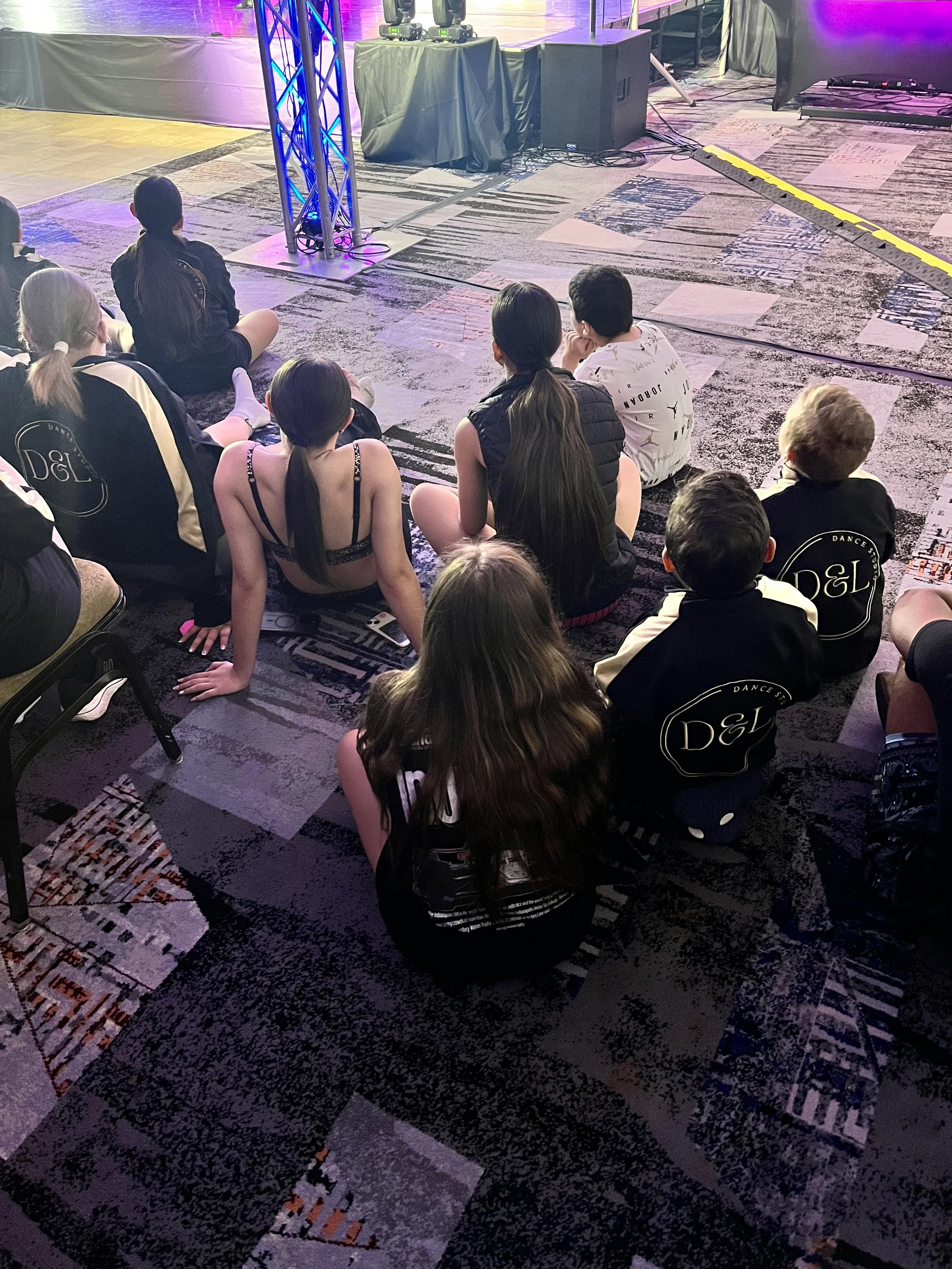 A group of young dancers sitting on a patterned carpet at the edge of a stage, watching a performance or event with stage lighting and equipment visible.