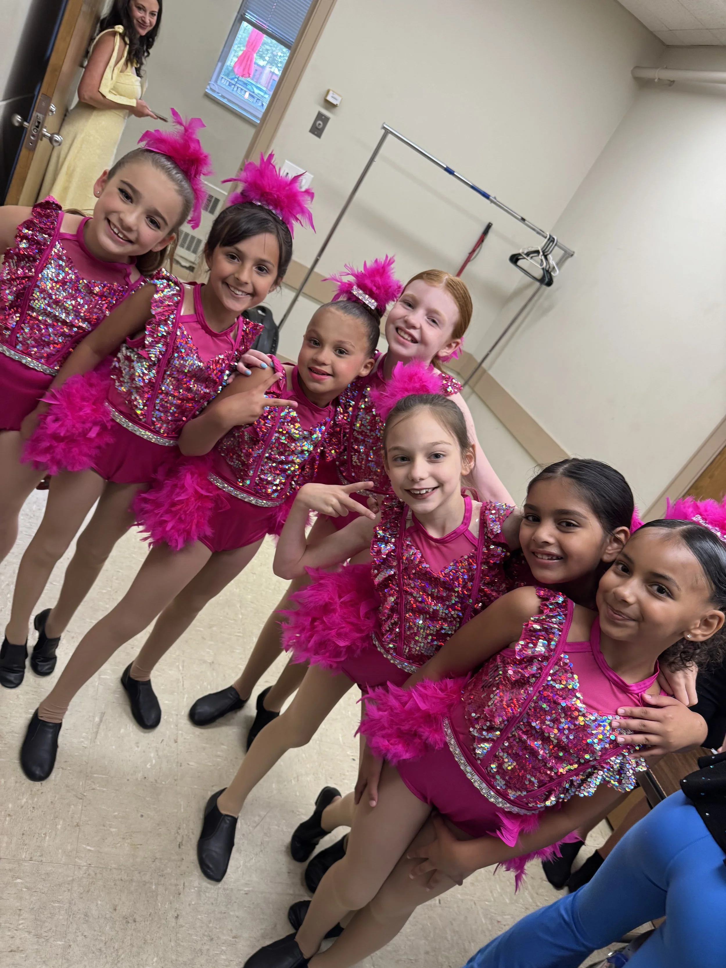 A group of young girls dressed in pink and sequined dance costumes, smiling and posing together backstage, with and adult woman standing near a door in the background.