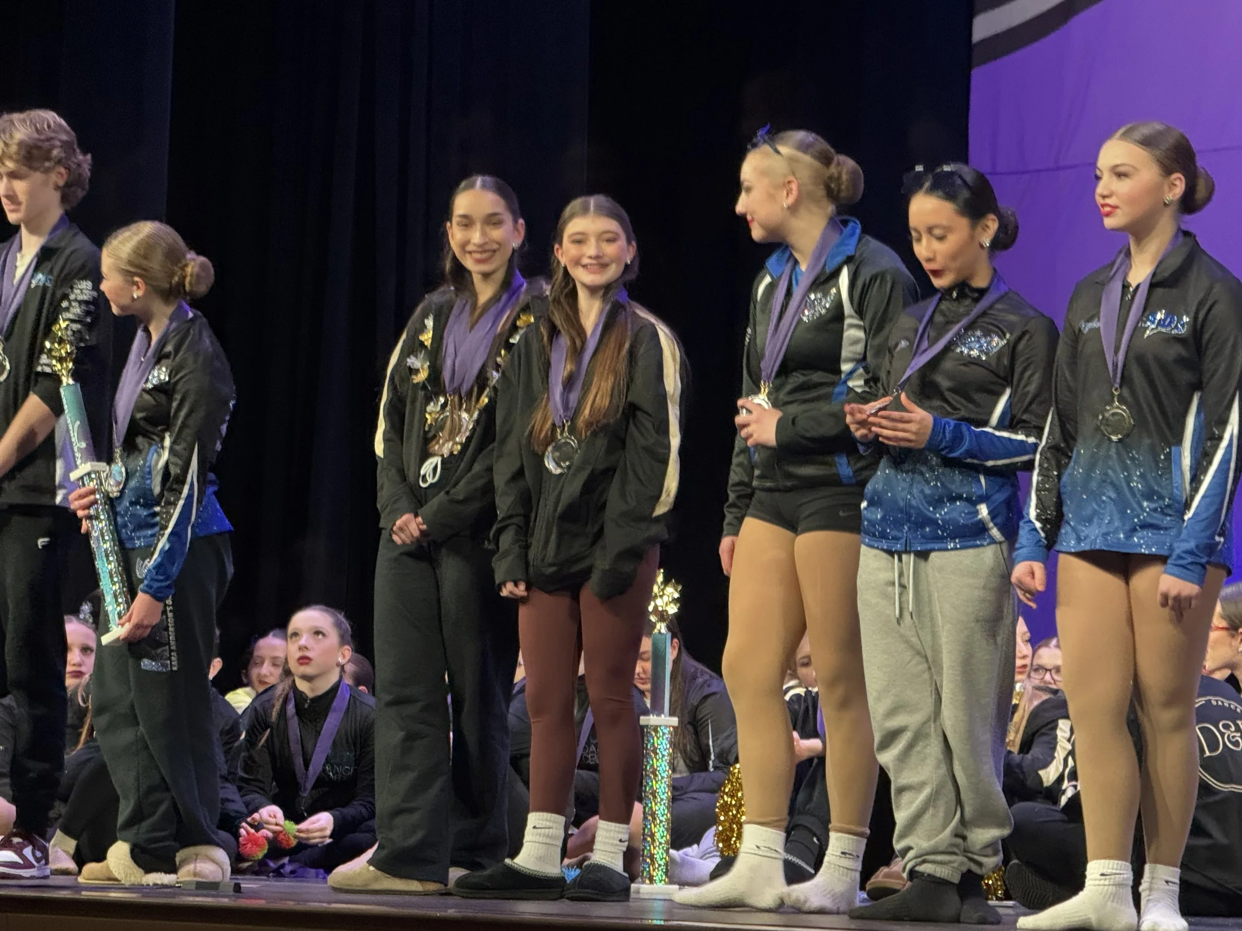 Group of young female figure skaters standing on stage with medals around their necks after a competition, some holding trophies.