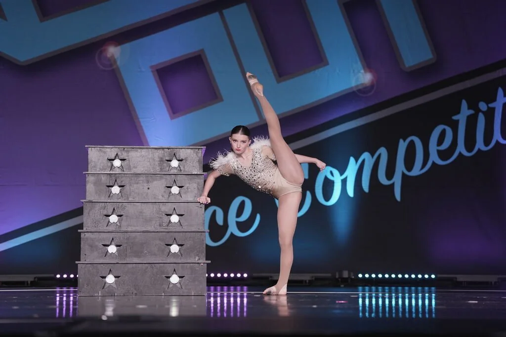 A young female dancer in a tan dance costume performing a high kick on stage at a dance competition. She is balancing on her left leg, with a large stack of gray wooden drawers beside her on stage, and a dark backdrop with the words 'Dance Competitio