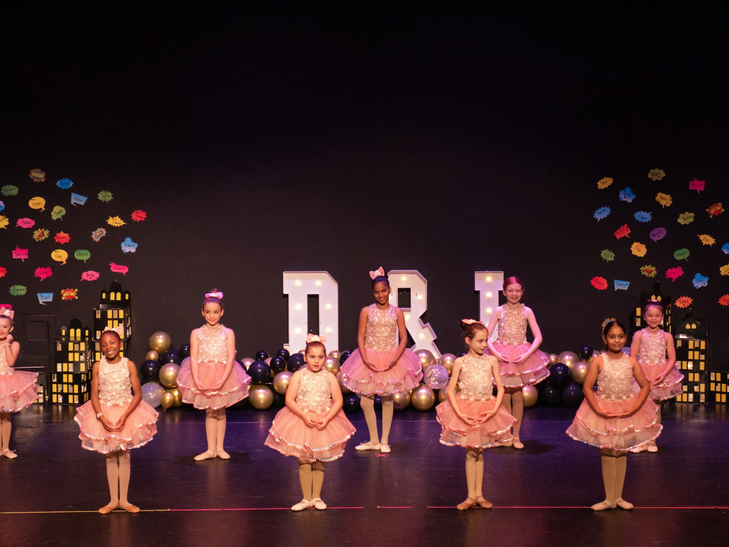Children in pink ballet costumes performing on stage with large illuminated letters and colorful wall decorations.