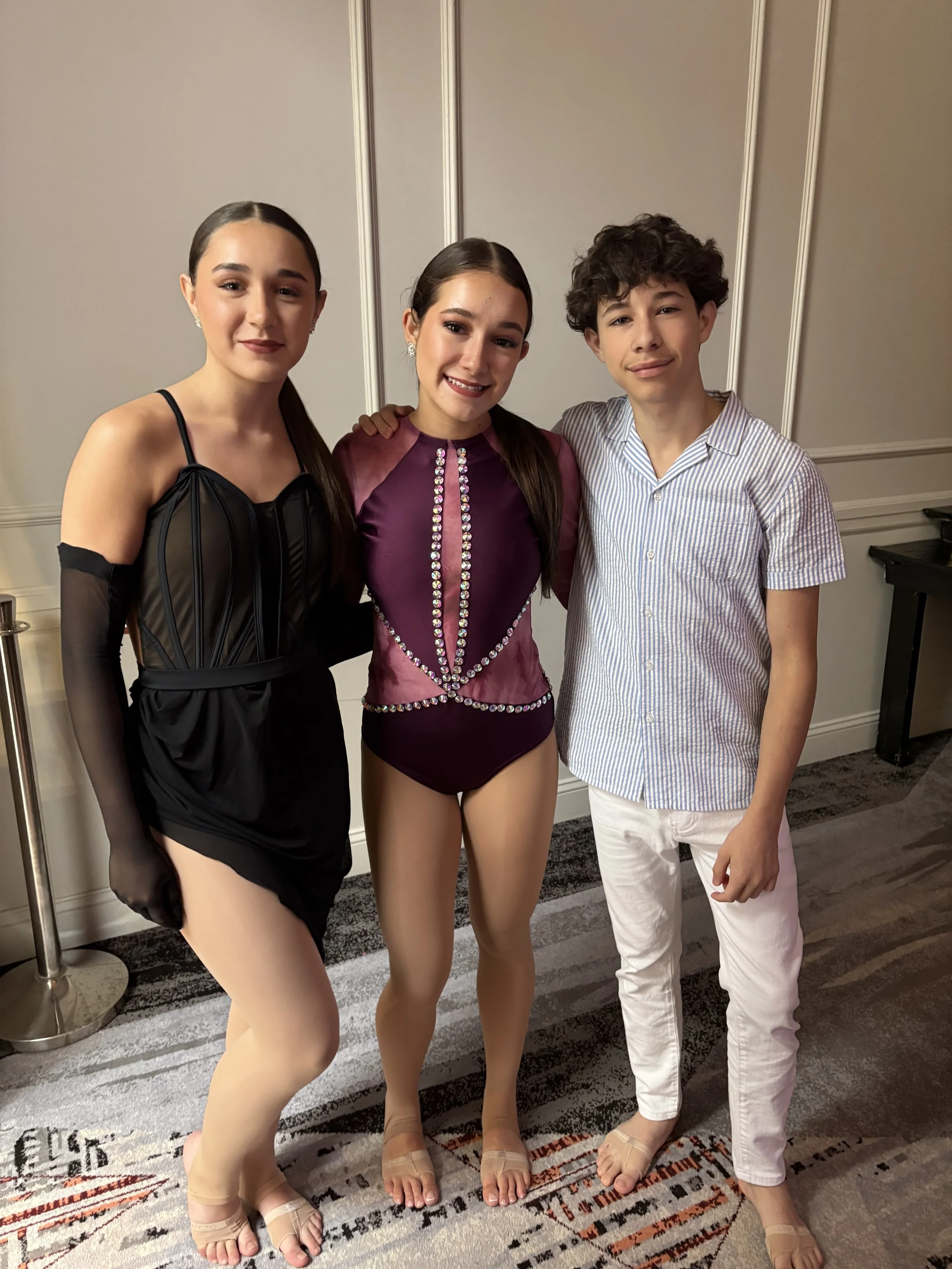 Three young individuals standing together in a room, dressed for a dance or competition, smiling at the camera.