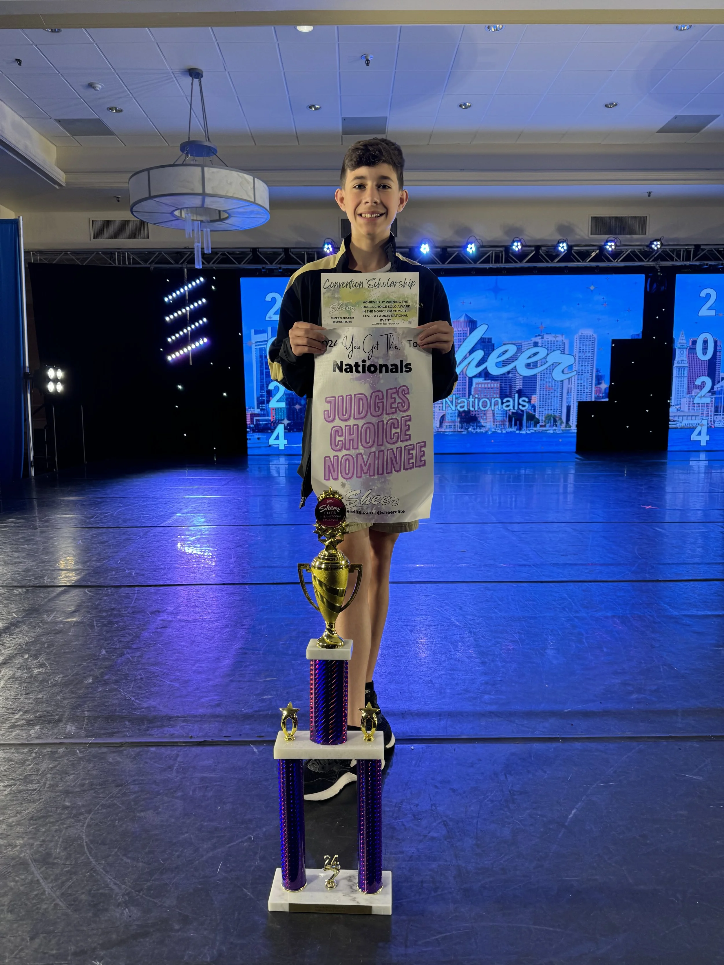 Young male cheerleader standing on a championship stage behind a large trophy, holding a certificate that reads 'Judges Choice Nominee.' The background features a large screen displaying 'Cheer Nationals' and the year 2024.