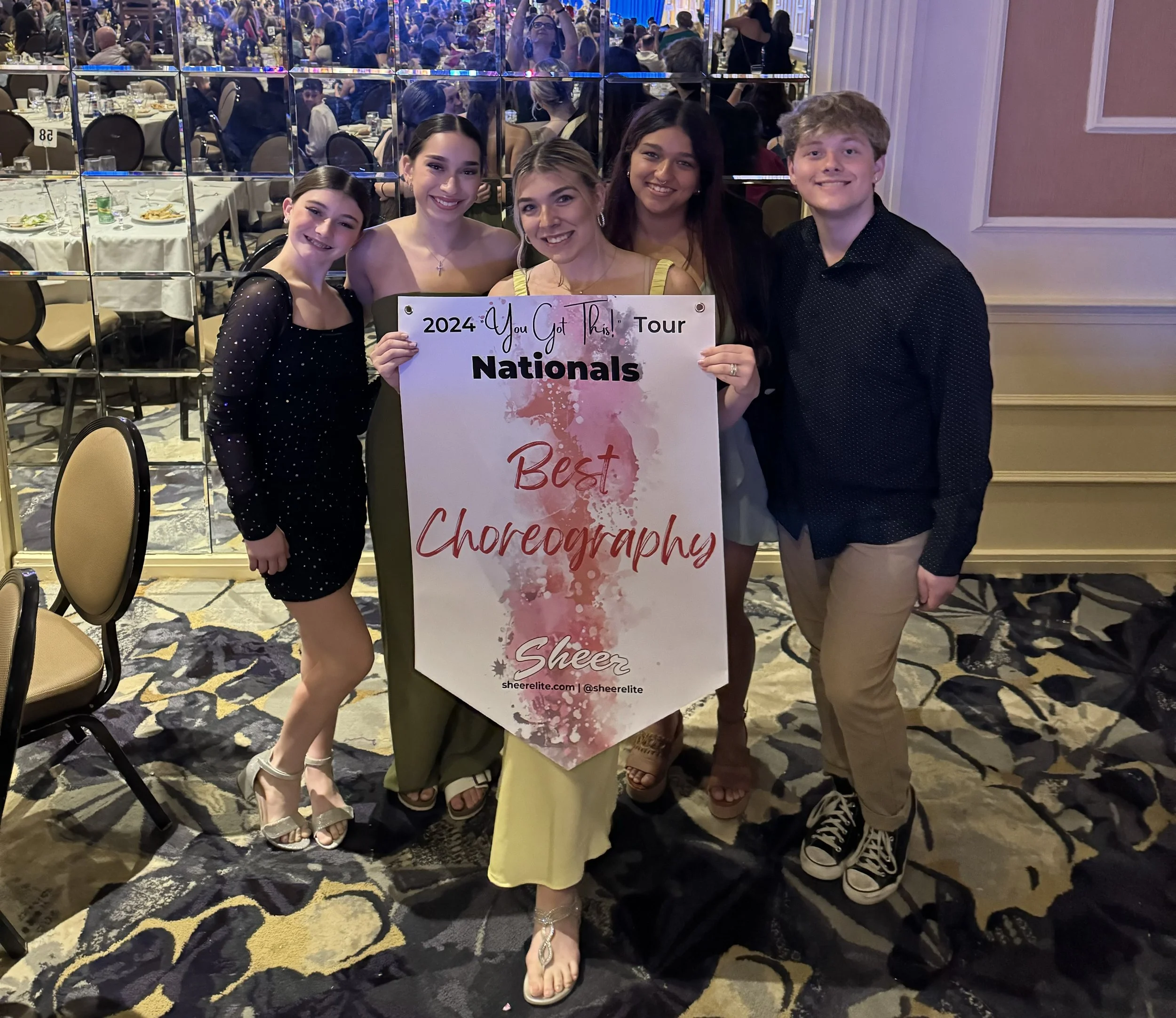 Group of five young people holding a large sign at an indoor event, with tables set for a celebration in the background.
