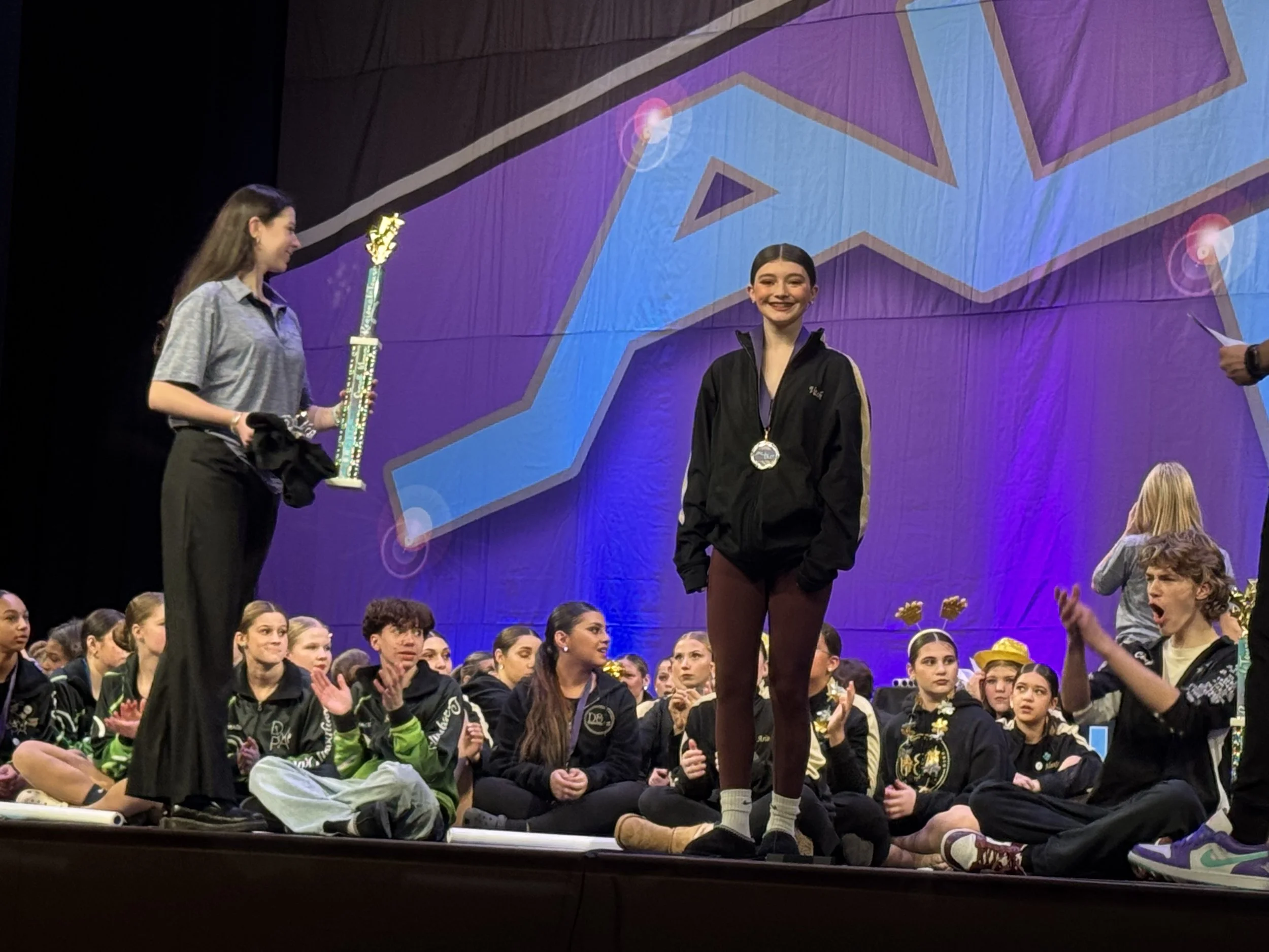 Young girl receiving an award on stage at a competition, surrounded by seated young girls, with a large colorful backdrop behind him.