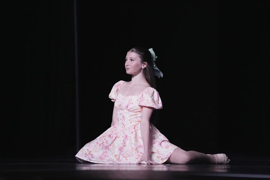 A young girl in a pink floral dress performing a split on a dark stage with a black background.