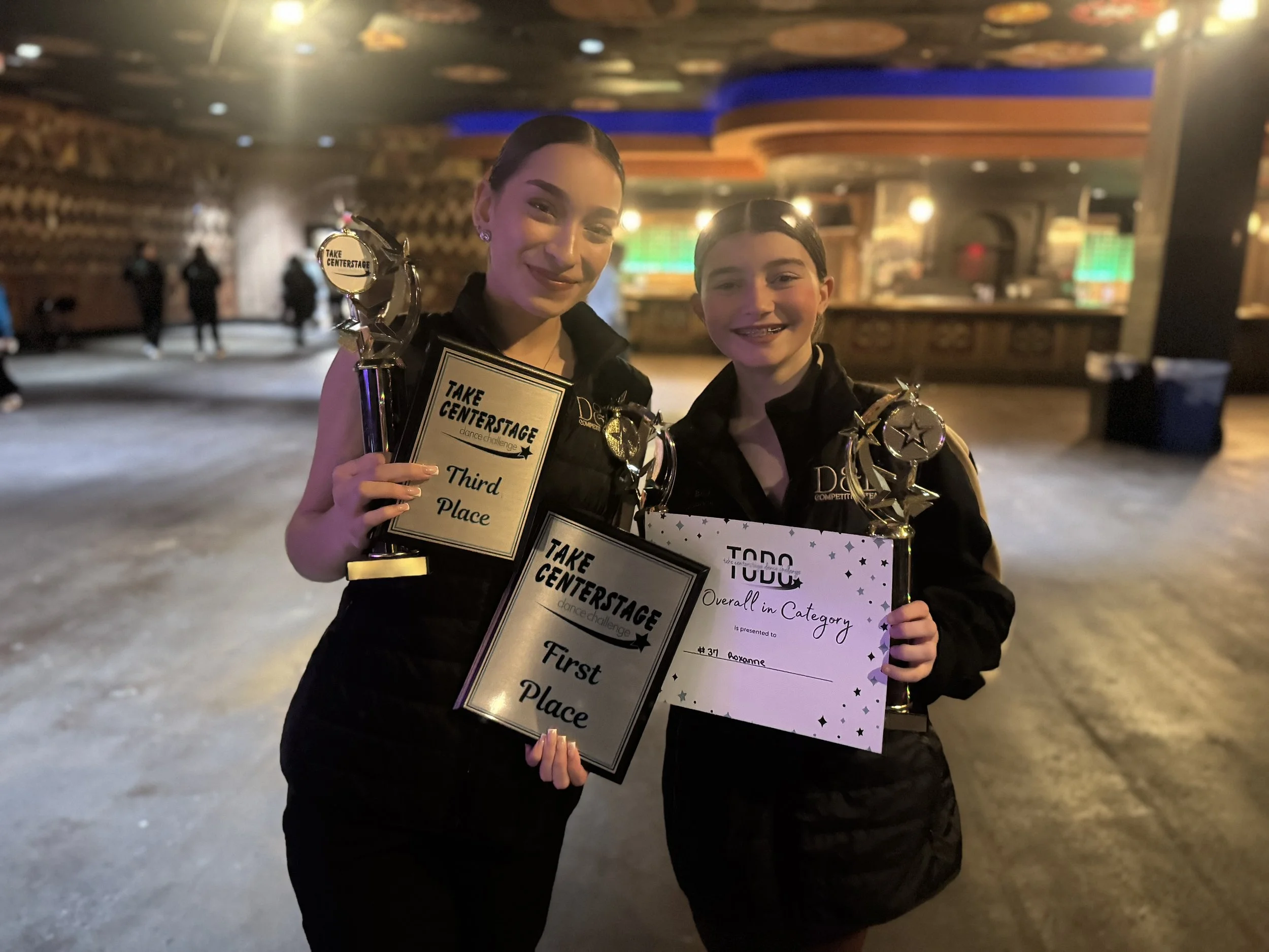 Two young girls holding trophies and certificates at a dance competition, standing in a dimly lit area with a bar in the background.