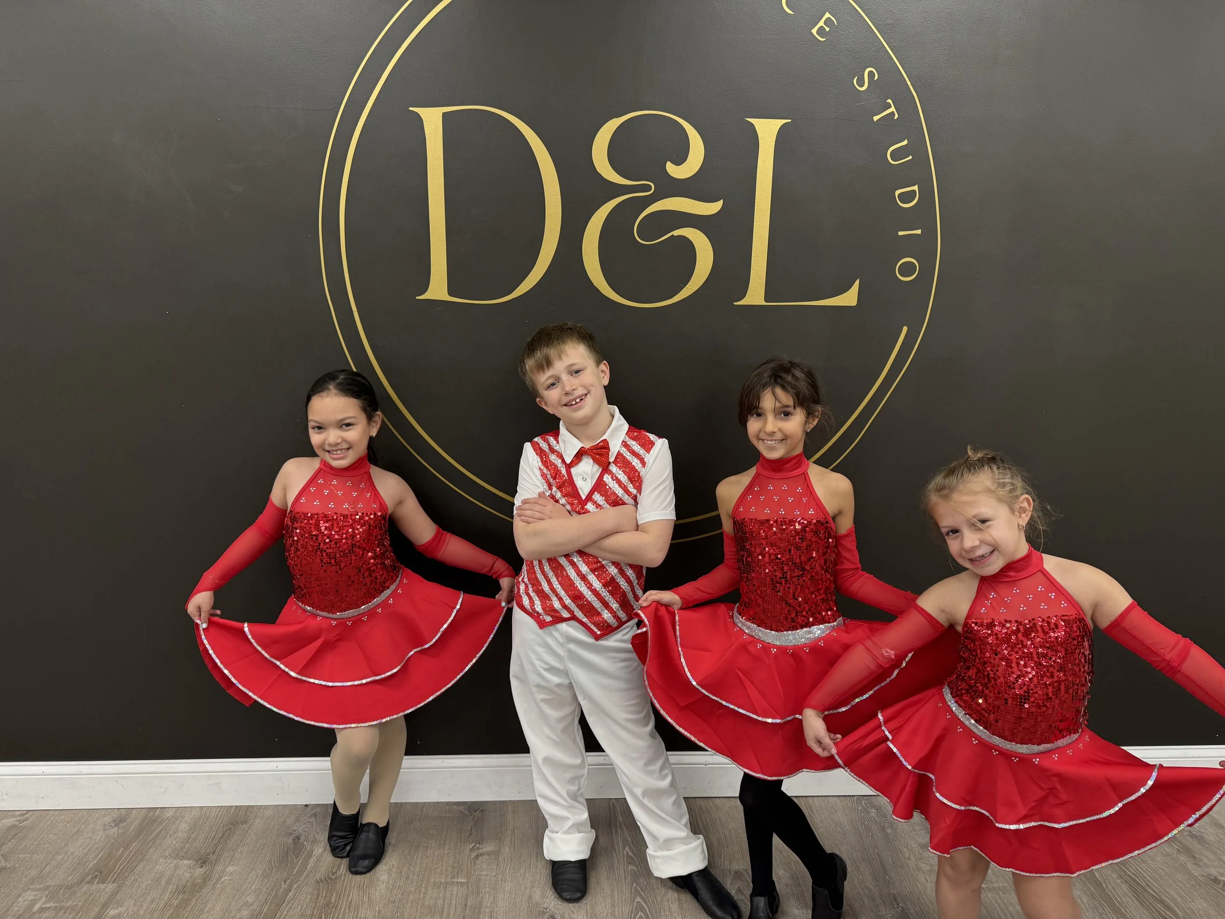 Four children dressed in dance costumes standing in front of a black wall with gold text that reads 'D&L' and 'Dance Studio'. The three girls are wearing red costumes with sequins and red skirts, and the boy is dressed in a white shirt with red and white striped vest and white pants.