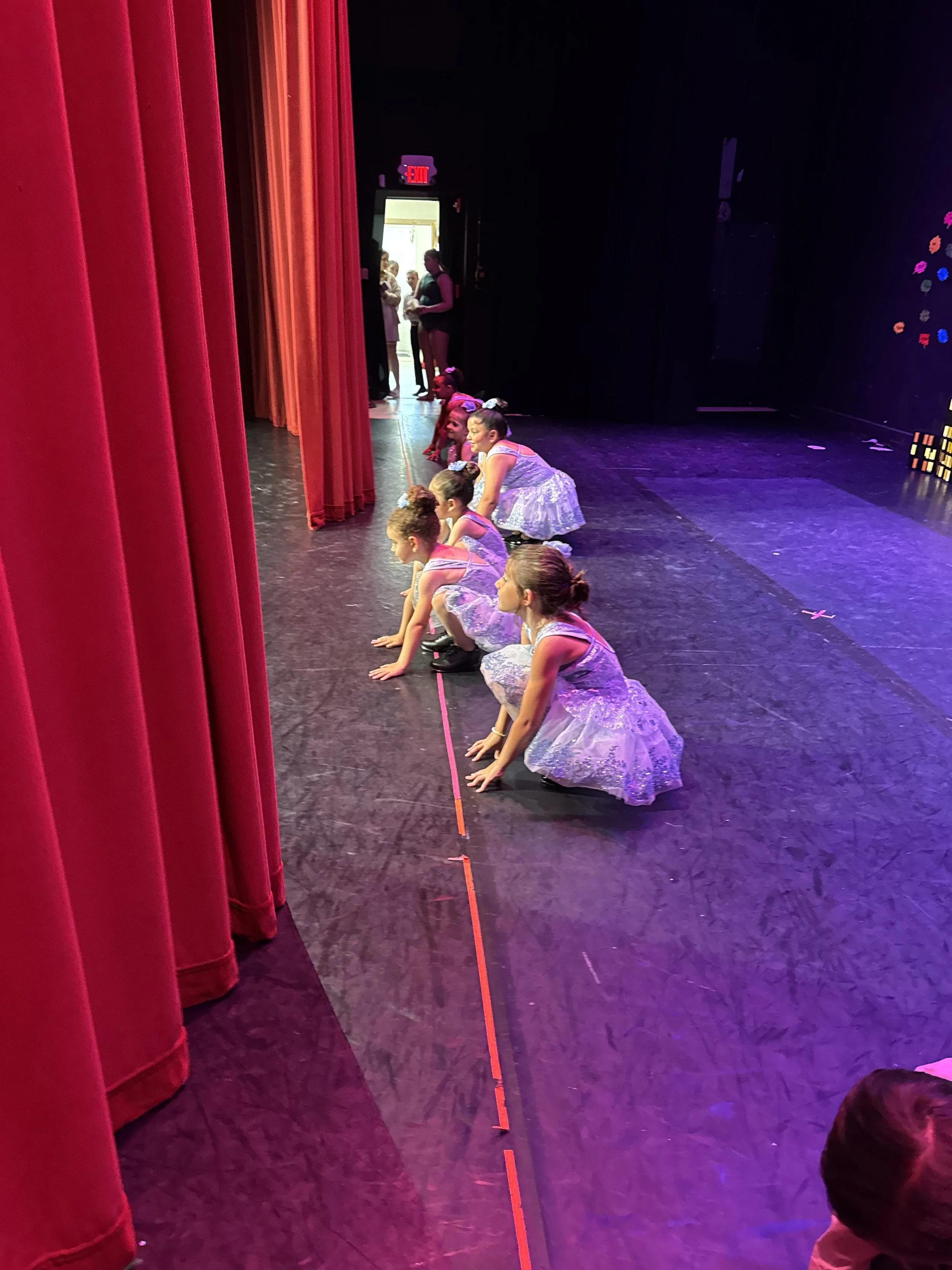 Young girls in ballet dresses preparing backstage before a dance performance, kneeling on the stage floor near the red curtains.