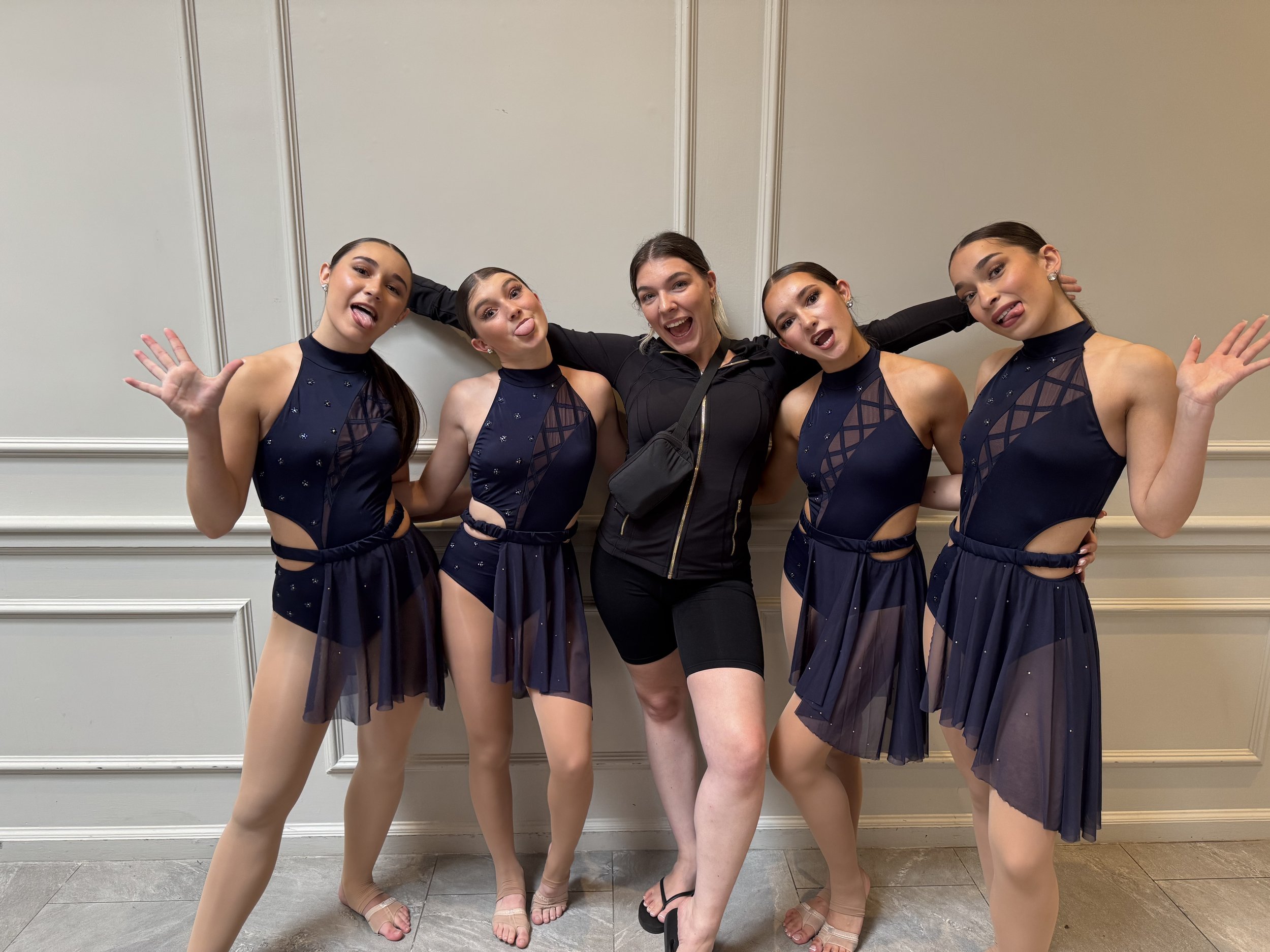 Five young women in navy blue dance costumes and a woman in black casual outfits posing together indoors against a plain white wall with paneled design.