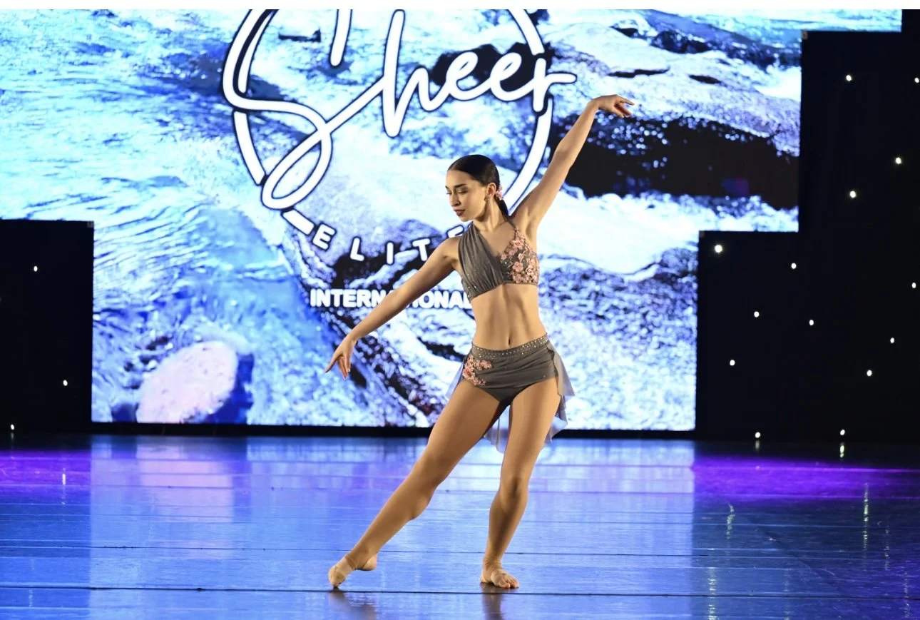 A young woman dancing on stage in front of a large blue and white background with the words "Shcek Elite International".