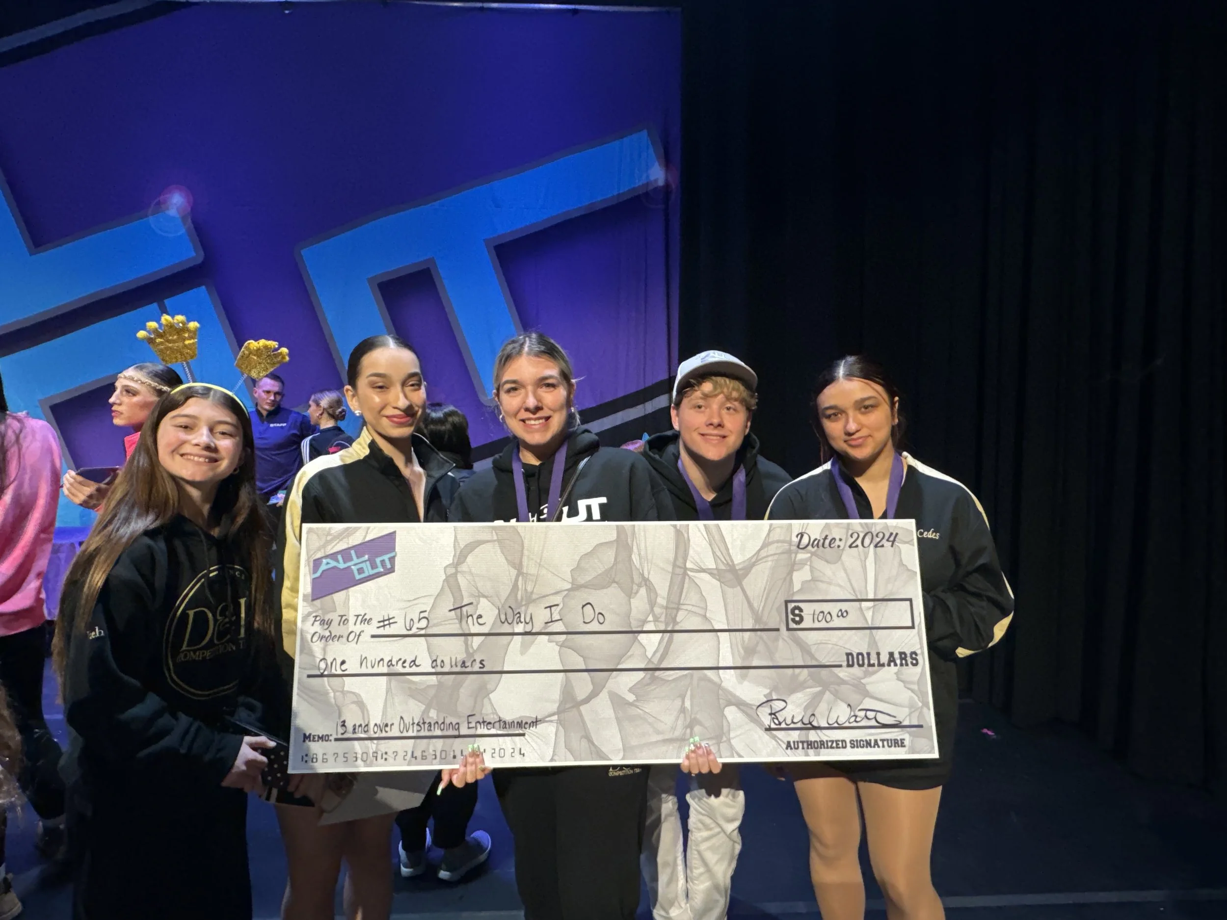 Group of five young people on stage holding a large ceremonial check, smiling at the camera, with other performers in the background wearing crowns and costumes.