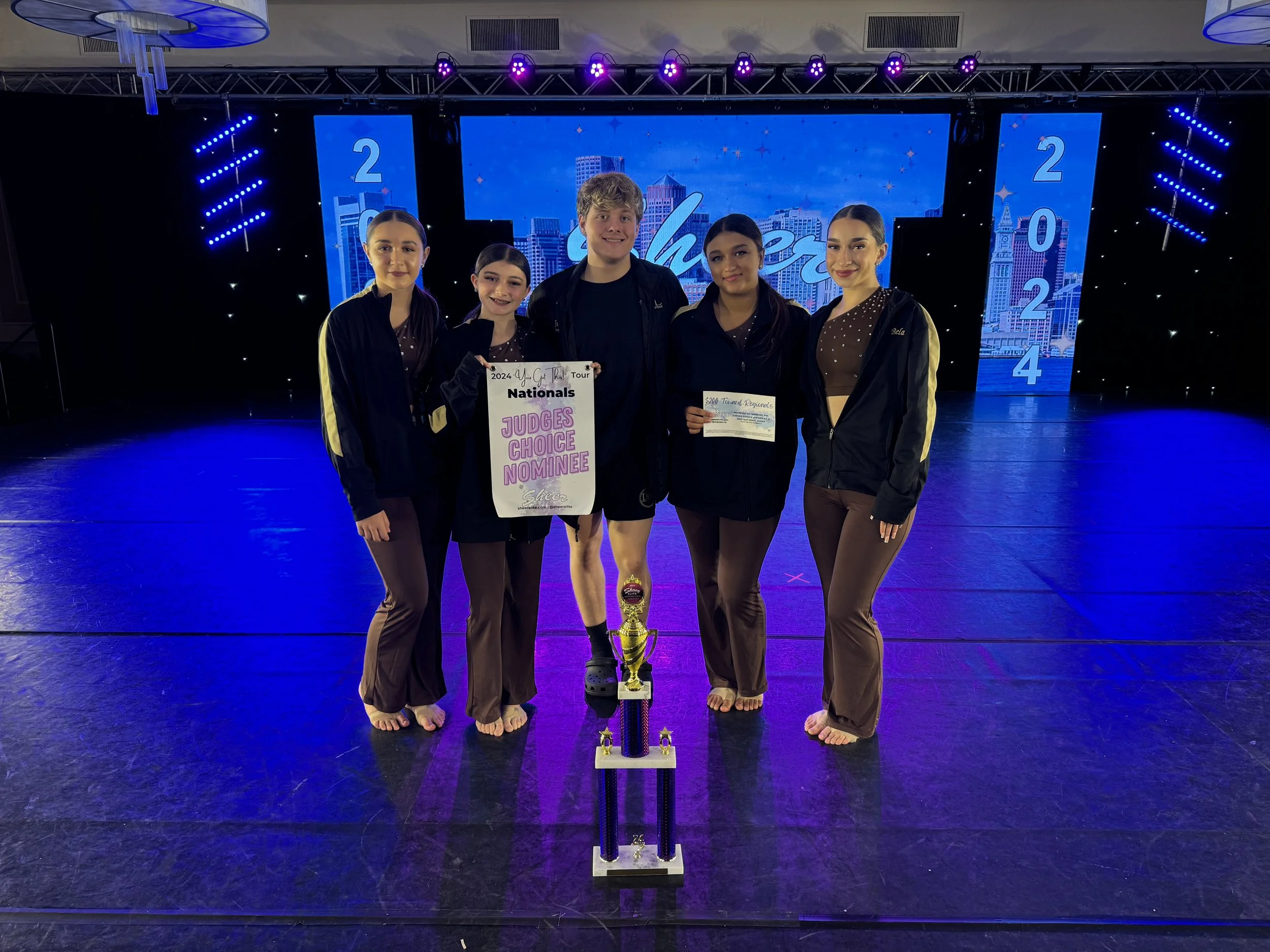 Group of five young dancers in black and brown dance costumes standing on stage with a large trophy, certificates, and a sign that reads "Judges Choice Nominee." Background displays blue digital screen with cityscape and the years 2022 and 2024.