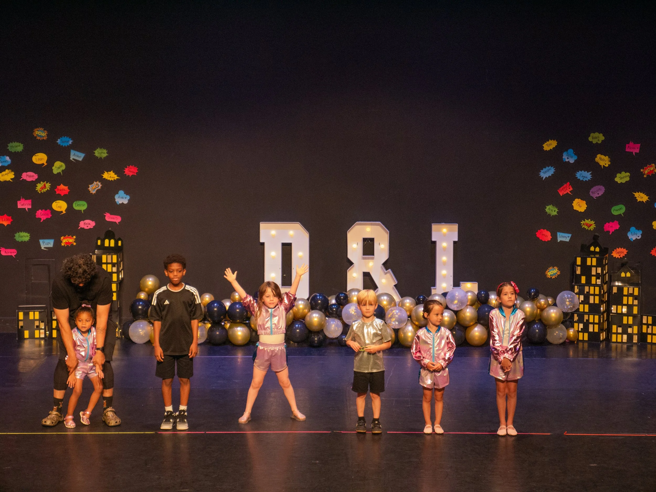 Children and an adult on stage with decorative balloons and large illuminated letters 'D' and 'U', in a theater setting with colorful speech bubble cutouts on the background wall.